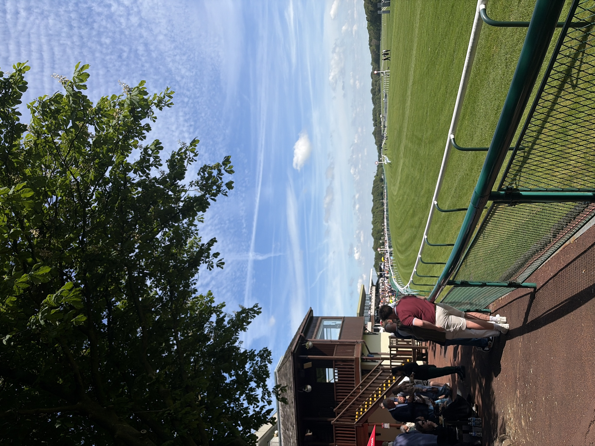 The image captures a daytime scene, likely at a sporting event, possibly a horse race. The location is presumed to be in the city of Unknown, Unknown. A crowd of people is visible along a fence line, some looking out onto a grassy field, while others sit in the shade of a wooden structure with steps. An individual in a maroon shirt and light-colored shorts is leaning against the fence. A large tree with green foliage is present in the left side of the frame. The sky above shows a partly cloudy, blue sky with contrails. A green, fenced barrier separates the viewers from the grassy field. The lighting suggests it is a sunny day.