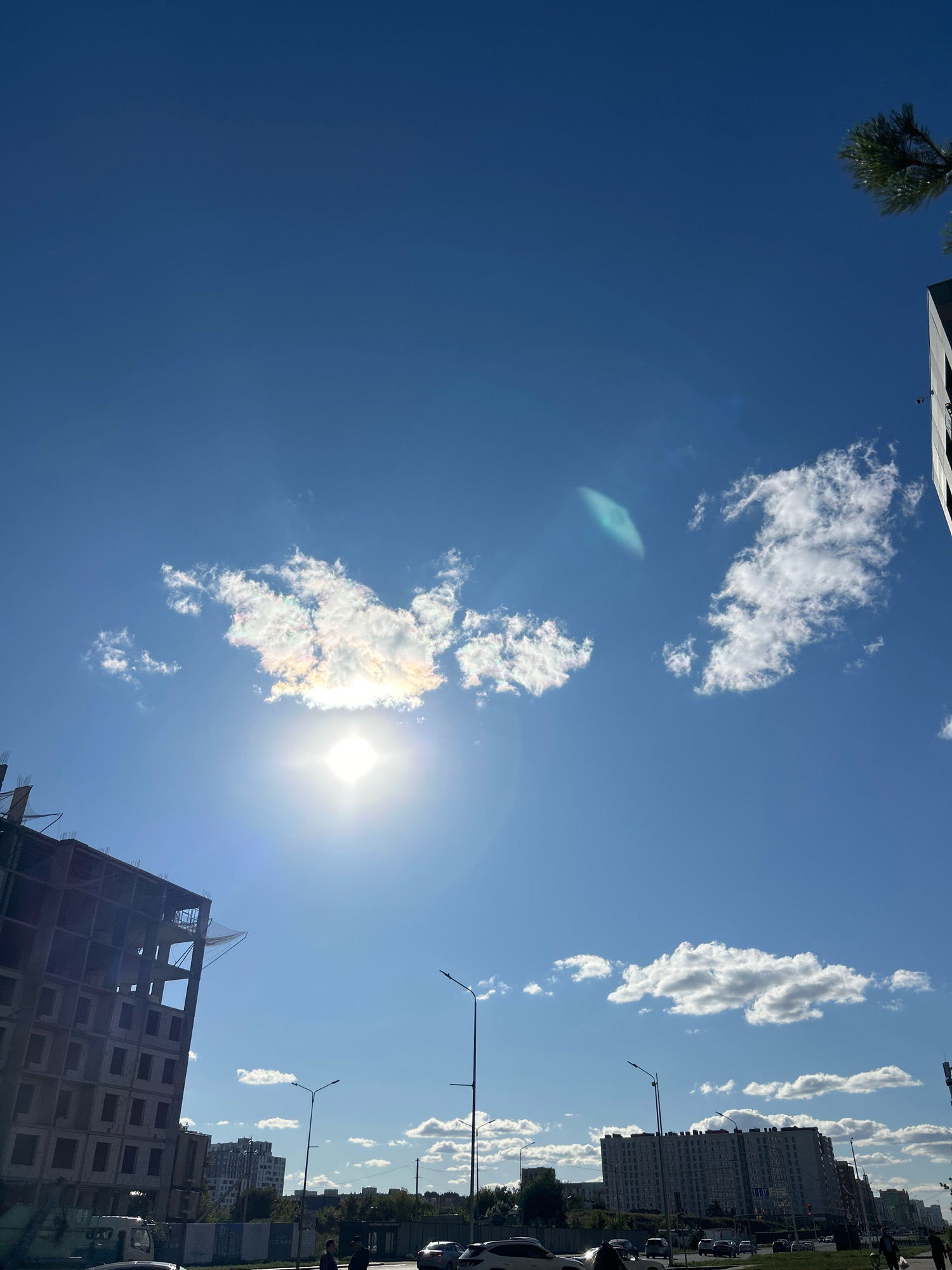 The image captures a bright, sunny day in Astana, Kazakhstan. The sky is a vibrant blue with scattered white clouds, one of which is illuminated by the sun, showing iridescent colors. In the foreground, a multi-story building under construction dominates the left side of the frame, its concrete structure partially visible. Several other residential buildings, appearing to be apartment blocks, are visible in the mid-ground. Streetlights are spaced along a road where several cars are driving and parked. A few individuals are walking on the sidewalk. The scene suggests an urban environment with ongoing development.