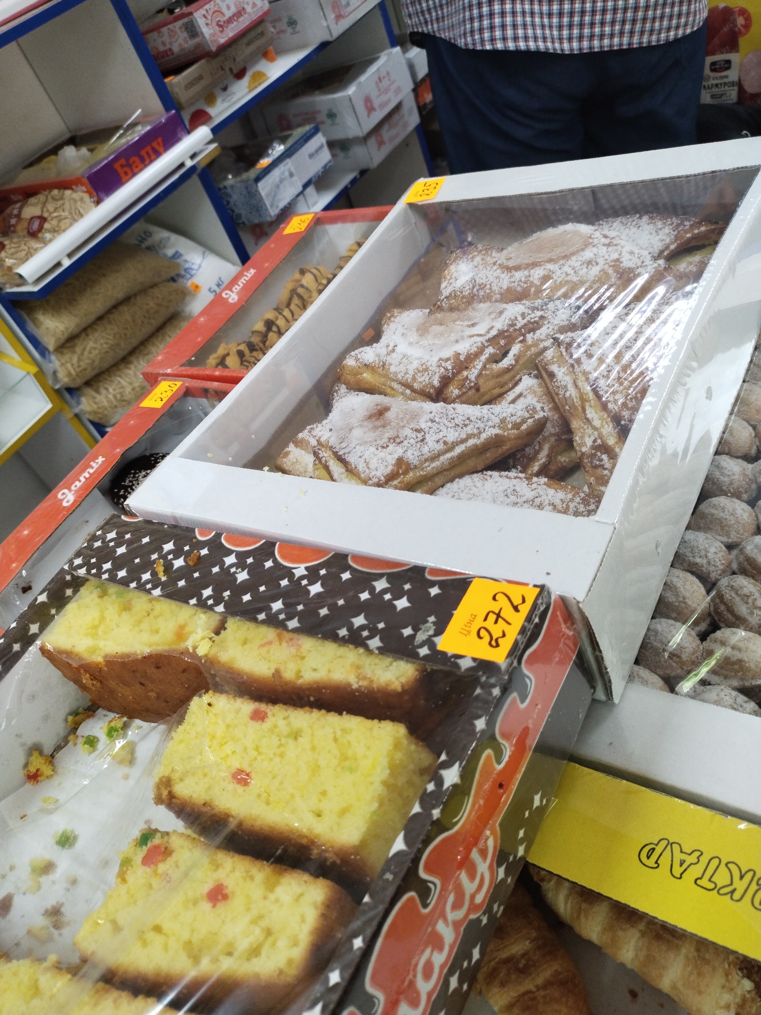 This is a close-up, angled photograph of a retail display of baked goods in Horishni Plavni, Ukraine. The products are arranged on shelves, with several items visible in clear plastic packaging. In the foreground, there are slices of yellow cake with colorful candied fruit pieces. To the right of the cake, a box contains round, cookie-like pastries covered in powdered sugar. Above these, another box holds larger pastries dusted with powdered sugar, possibly strudels or turnovers. The background shows more shelves with packaged food items, including what appears to be pasta or noodles in plastic bags and boxes with Cyrillic labeling. A yellow price tag with the number "272" is visible on one of the pastry boxes. The lighting suggests an indoor environment, likely a grocery store or bakery. A person's torso is partially visible at the top, wearing a plaid shirt and dark blue pants, indicating the scene is during business hours.