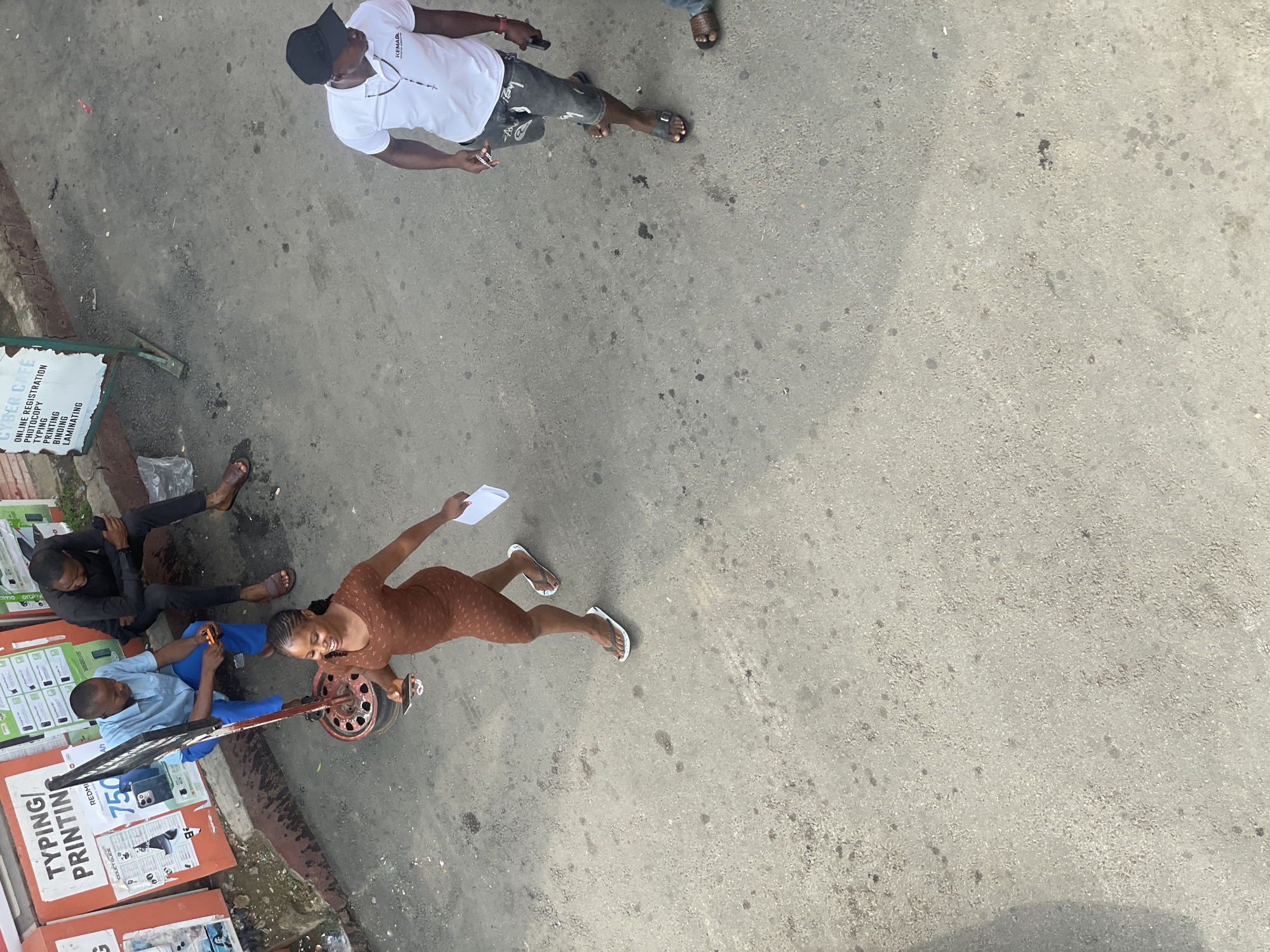 The image captures a street scene in Abuja, Nigeria, with a focus on pedestrian activity and commercial signage. In the foreground, a woman in a brown patterned dress and white flip-flops walks away from the camera, holding a smartphone and a piece of white paper. To her right, a man wearing a white t-shirt, dark shorts with white lettering, and a black baseball cap walks towards the camera, holding a smartphone in his right hand. His t-shirt displays the logo "KEMAD".
In the background, two men are seated near a street stall or shop. One man, wearing a light blue shirt and dark blue trousers, is seated on the ground and looking at his phone. Beside him, a man in a black shirt and dark trousers is also seated and appears to be engaged with his phone. Behind them, a sign reads "TYPING/PRINTING" and displays a price of "750", suggesting a business offering these services. Another sign with green text on a white background, positioned to the left and behind the first sign, lists services like "CYBER CAFE", "ONLINE REGISTRATION", "PHOTOCOPY", "TYPING", "PRINTING", "BINDING", and "LAMINATING". The ground is paved with asphalt, showing some wear and tear with dark stains and lighter patches. The overall impression is of a bustling urban environment with visible commerce and everyday life unfolding on the street.