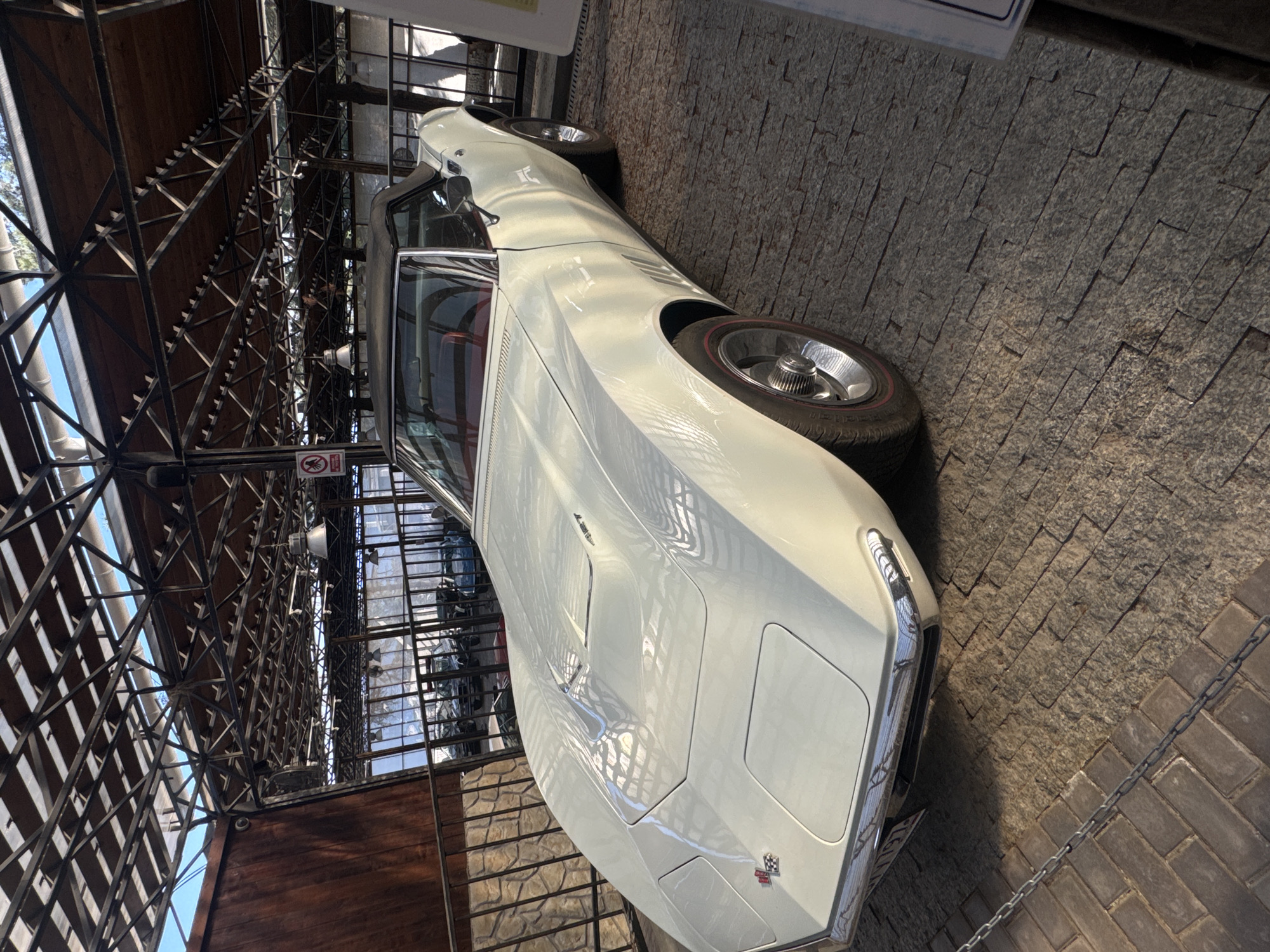 The image captures a white vintage Chevrolet Corvette parked indoors on a brick surface. The car is angled towards the viewer, showcasing its front design, including the headlights, emblem, and tire. The background includes architectural details of a structure with metal beams and glass windows, suggestive of an indoor showroom or museum setting. The location is город Ялта, Ukraine.