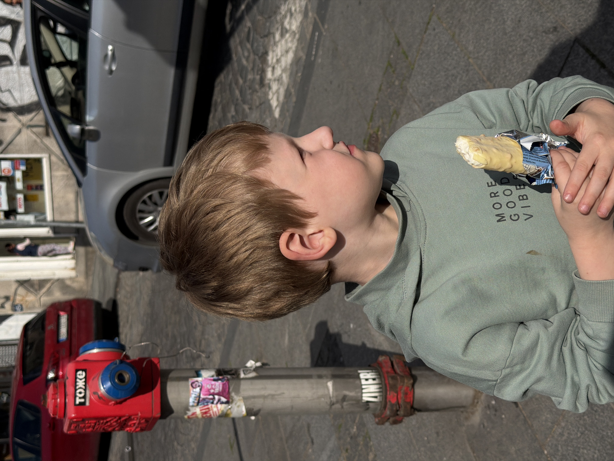 This image depicts a young boy engaged in a casual activity on a sunny city street, identified as Belgrade, Serbia. The boy, appearing to be around 5-8 years old with light brown hair, is shown from the chest up, looking upward and slightly to his right with a calm and possibly curious expression. He is wearing a light green long-sleeved sweatshirt that reads "MORE GOOD VIBES" on the front. In his hands, he holds a partially unwrapped white chocolate-coated ice cream bar.
The scene unfolds in bright daylight, indicated by strong shadows cast on the paved ground, suggesting a clear and sunny day. In the background, a light gray compact car is parked on what appears to be a cobblestone and asphalt street. To the lower left, a distinctive red fire hydrant, uniquely stylized to resemble a retro camera, is visible with the Cyrillic text "TOXKE" inscribed on its side. A utility pole with various stickers, including a partially visible one that reads "ZINER!", stands nearby. Further back, elements of an urban environment such as graffiti on a wall and a small white display case can be seen, along with the lower legs of another person in the distance. The overall impression is a vibrant urban setting where everyday moments of enjoyment take place.