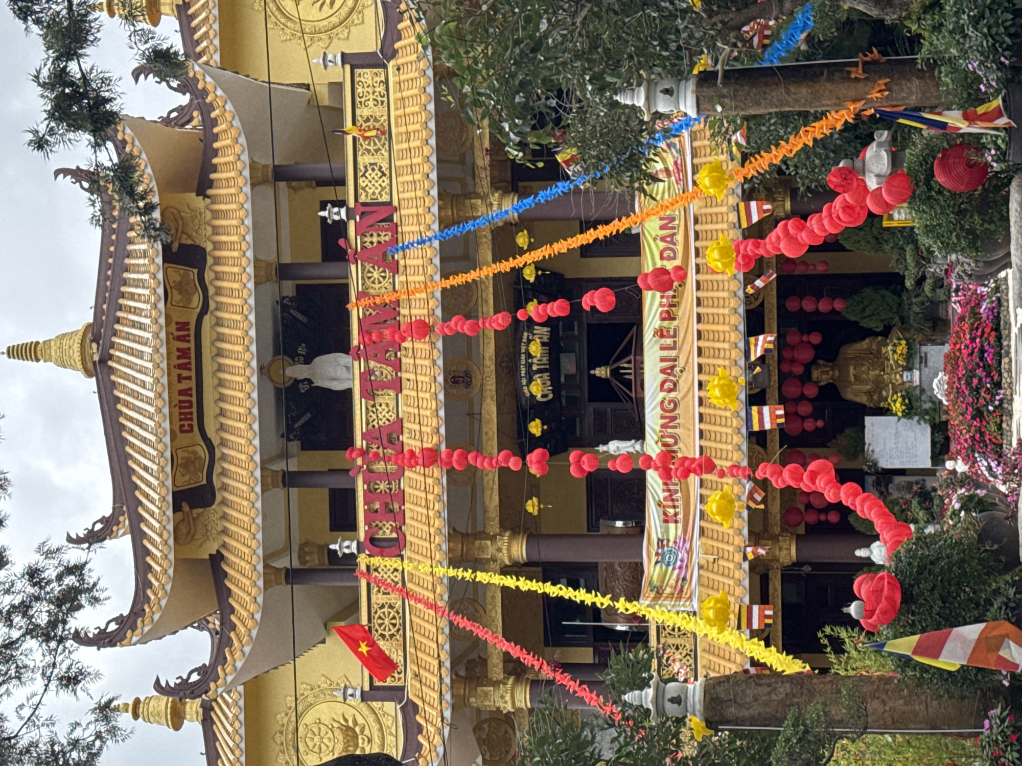 The image shows the exterior of Chua Tam An, a Buddhist temple in Da Lat, Vietnam. The temple is decorated with colorful garlands of red and yellow paper lanterns, as well as red and blue streamers. The temple itself is a large, multi-tiered structure with a golden roof and ornate architectural details. In front of the temple, there are statues of Buddha and other religious figures, as well as a large display of flowers. The overall scene suggests a festive or celebratory occasion. The temple is located on a hillside, with trees and vegetation visible in the foreground and background.