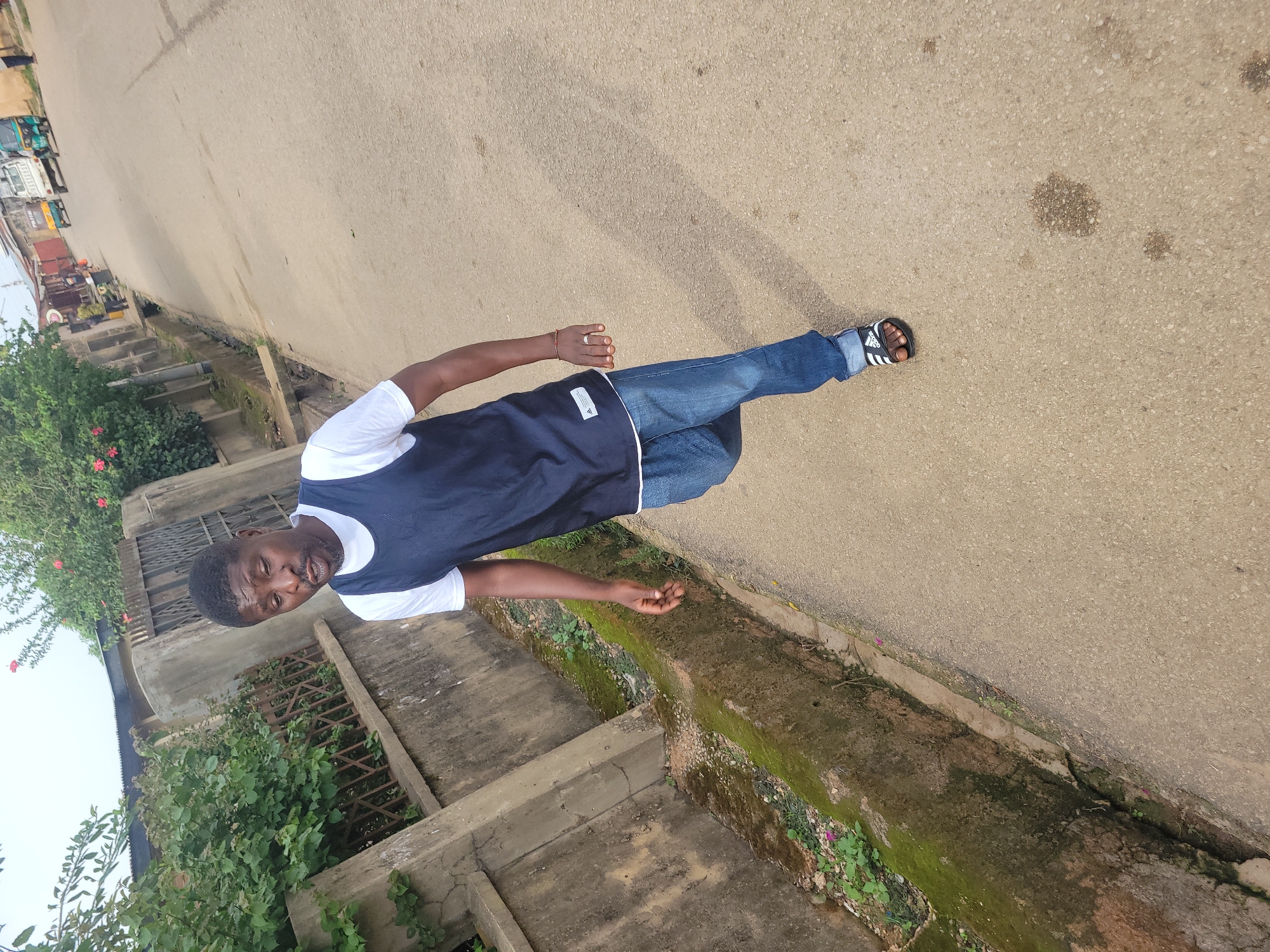 A Black man is walking on an asphalt road in Jos, Nigeria. He is wearing a white t-shirt, a navy blue sleeveless vest, blue jeans, and black Adidas slides. The man is looking down and to his left with his mouth slightly open. To his left is a concrete drainage ditch filled with moss and sparse vegetation. Behind the ditch is a concrete wall with a metal fence and dense green bushes. In the background, on the right side of the road, there are buildings, some trees, and vehicles, including a white truck and a green tricycle. The sky is overcast.