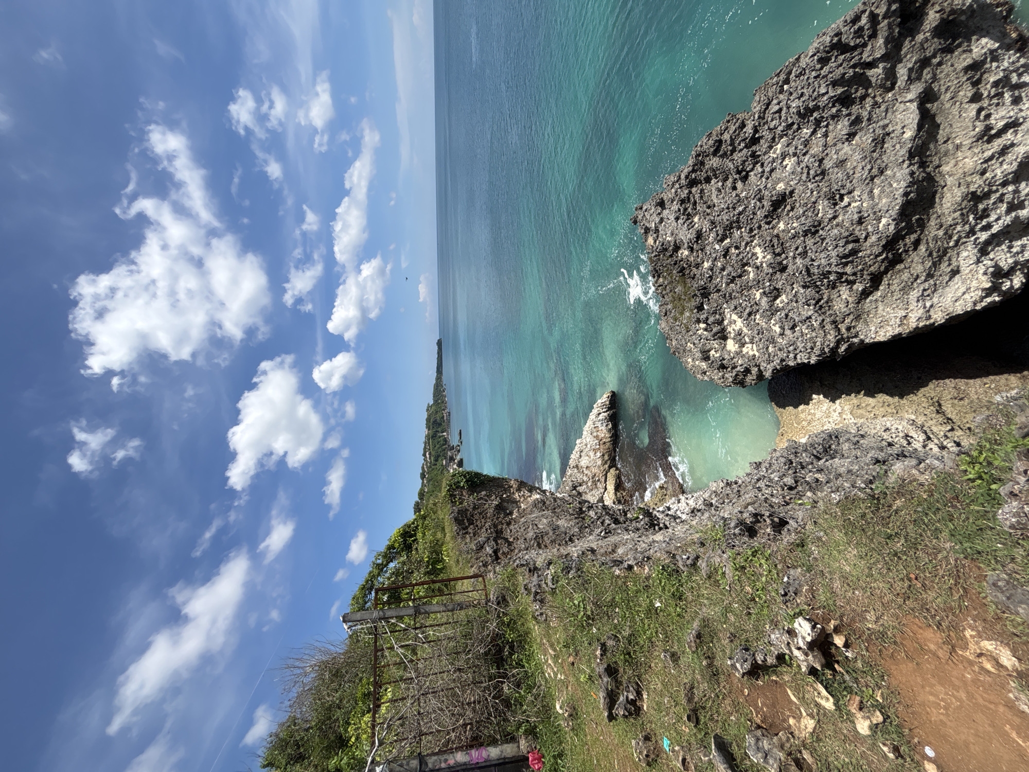 The image shows a coastal scene. A rocky cliff dominates the foreground with vegetation growing on its surface. Beyond the cliff, the turquoise-colored ocean stretches out. The sky is blue with scattered white clouds. In the background, a strip of land can be seen along the coast. There is a rusted metal structure on the side of the cliff, and a bit of trash.
Location: Unknown, Indonesia.