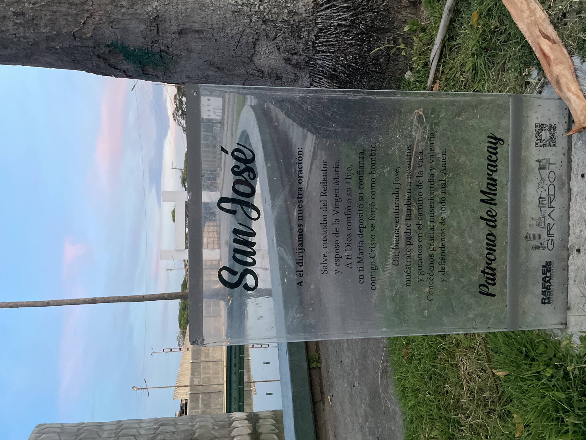 The image shows a glass plaque with text in Spanish, placed outdoors next to a tree trunk and some grass. The plaque is positioned in front of a body of water, possibly a fountain or pond, with a concrete structure in the background. The sky above is a blend of blue and pink, indicating it might be dusk or dawn. The text on the plaque appears to be a prayer dedicated to "San José" (Saint Joseph), with the bottom of the plaque indicating "Patrono de Maracay" (Patron of Maracay), suggesting the location is the city of Maracay, Venezuela. The lower part of the plaque also contains a QR code and logos. There are no people visible in the image.