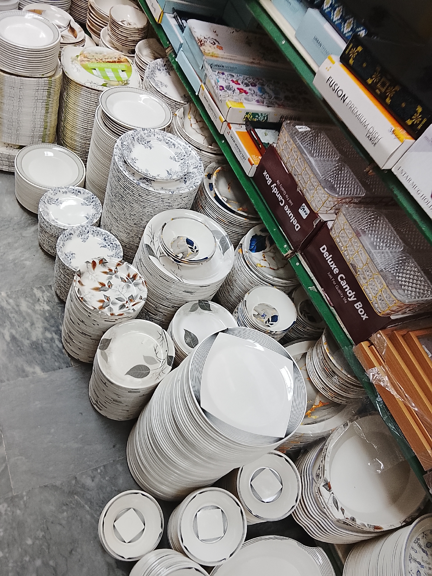 The image shows a retail display of dinnerware, likely within a store in Gulgasht Colony, Pakistan. Stacks of various plates and bowls, predominantly white with diverse patterns (floral, leaf, geometric), occupy the floor and shelf space. Some items are in original packaging with brand names "URBAN TRENDS" and "FUSION PREMIUM DISH". Brown boxes labelled "Deluxe Candy Box" are also on the shelf. The floor appears to be grey tiled.