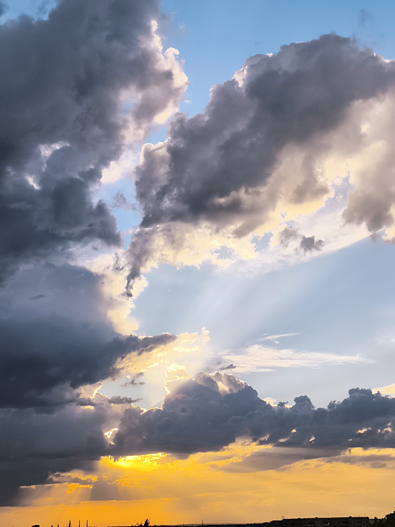 The image captures a dramatic sky with a mix of dark, heavy clouds and bright, sunlit patches. The sun is low in the sky, casting golden and orange hues across the lower part of the frame, illuminating the undersides of the clouds with a warm glow. Rays of sunlight pierce through breaks in the clouds, creating visible beams that extend downwards. The upper portion of the sky is a lighter blue, with thinner, scattered clouds. In the distance, a dark, silhouetted line suggests a horizon, possibly with buildings or industrial structures, characteristic of an urban or industrial area like the city administration of Qarağandı city, Kazakhstan. The overall impression is of a dynamic and visually striking sky, possibly at sunset or sunrise, with storm clouds beginning to break. There are no people or specific actions depicted; the focus is entirely on the natural atmospheric conditions.