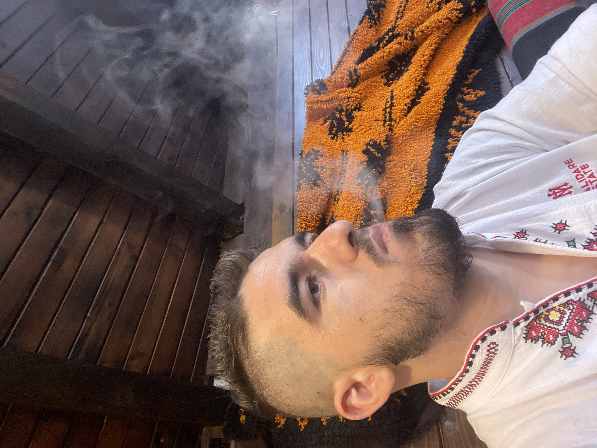 A man with a short, buzzed haircut and a beard exhales a plume of smoke. He is wearing a white shirt with red, black, and yellow embroidery around the collar and down the front. Behind him, an orange and black patterned textile is draped over a wooden surface. The ceiling is made of dark wooden planks. The man's face is turned to the right in profile.