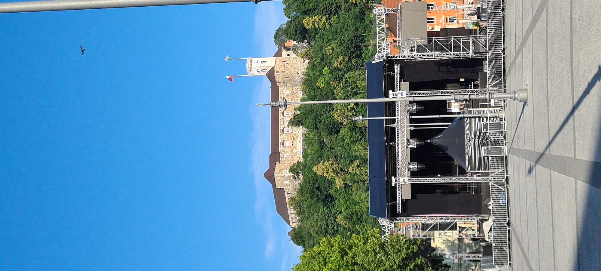 The image depicts an outdoor scene in Ljubljana, Slovenia, with a large stage structure in the foreground and Ljubljana Castle in the background. The stage is elaborate, constructed with metal scaffolding and draped with dark blue fabric. Several spotlights are visible, suggesting it is set up for an event, possibly a concert or festival. Behind the stage, lush green trees obscure much of the castle, but parts of the stone walls and a tower are visible. Two flags fly from the castle tower, one green and white, and another red, white, and blue. The sky is a clear, bright blue, indicating a sunny day, likely during daytime.
The scene appears to be a public square or plaza, with a paved ground marked by grey lines. A long, thin shadow stretches across the paving stones from an unseen object, likely a pole or structure. In the far left of the image, a small bird is visible in flight against the blue sky. The overall impression is one of anticipation for an event, with the historic castle serving as a backdrop to the modern stage setup. No people are prominently featured, and no visible text is present in the image.