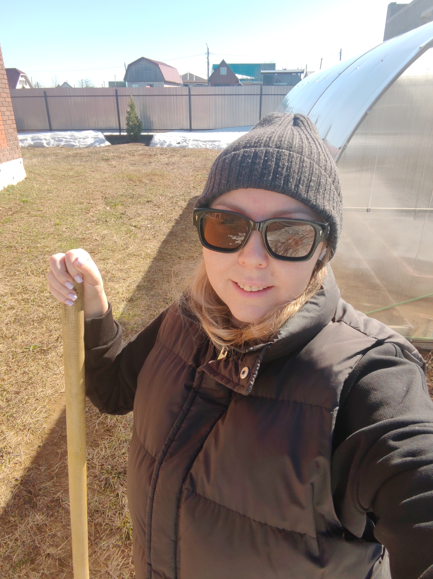 A woman stands outdoors on a sunny day in Chaikovsky, Russia. She is wearing a dark gray beanie, large black sunglasses, a black vest over a black hooded sweatshirt, and a white wireless earbud is visible in her left ear. She is holding a wooden-handled tool (likely a shovel or similar garden implement) in her left hand. The woman is facing the camera, and the photograph appears to be a selfie.
Behind the woman, a metal-framed greenhouse is visible to the right. In the background, there is a light-colored metal fence, a brick building, and a patch of melting snow. The ground is covered in dry grass and straw.