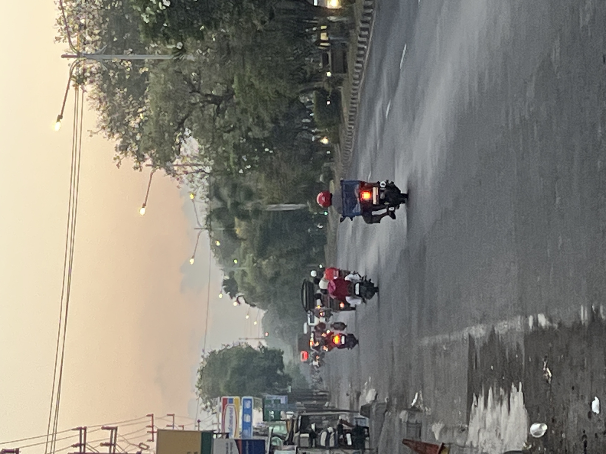 The image captures a street scene in Ngaban, Indonesia, during a time of low light, likely dawn or dusk, indicated by the soft, warm hues of the sky and the presence of streetlights. A two-lane asphalt road dominates the foreground, with a distinct curb and sidewalk on the left. The sidewalk appears to have some debris.
Several individuals on motorcycles are traversing the road. On the left side, a person in a black and white striped shirt is seated on a motorcycle parked near a building with various signs, one of which appears to be for "Indomaret". Further down the road, a convoy of motorcycles is visible, some carrying passengers. One motorcycle in the center has a rider in a red jacket, and another in the foreground is ridden by a person in a blue delivery box. The motorcyclists are wearing helmets, and some have casual attire.
The environment includes trees lining the right side of the road, with some streetlights casting a yellowish glow. In the background, more trees and buildings are discernible, hinting at an urban or semi-urban setting. A large truck is also present in the distance, among the other vehicles. The overall atmosphere suggests everyday commuting or delivery activities in a bustling Indonesian town.