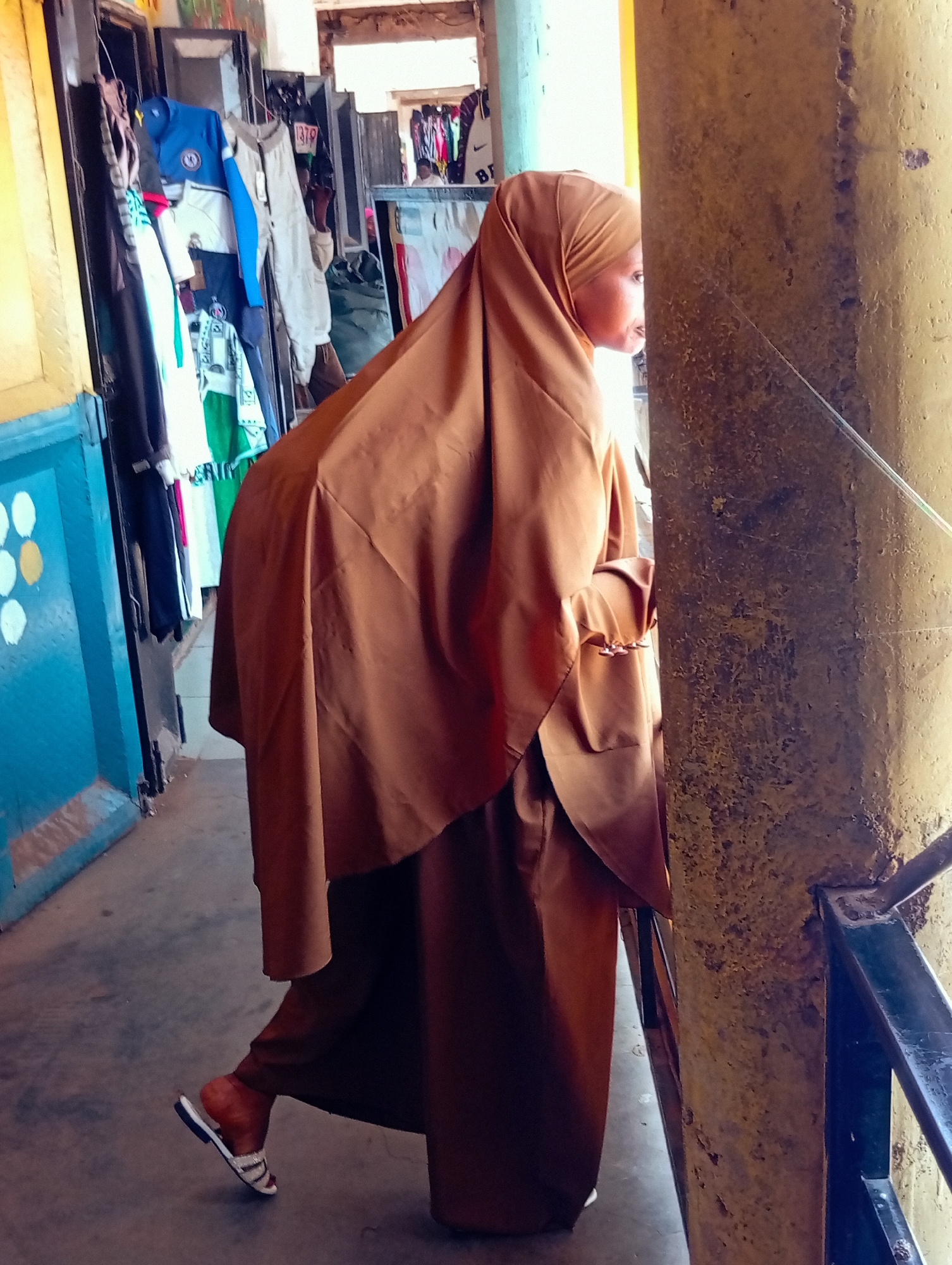 A person wearing a brown hijab and a long brown dress stands in an outdoor market in Bukuru, Nigeria. The person is looking to the right and is partially obscured by a yellow, rough-textured pillar. In the background, there are racks of clothing for sale, including t-shirts and jackets. The ground is paved. The person is wearing white sandals with a black sole and a decorative strap.