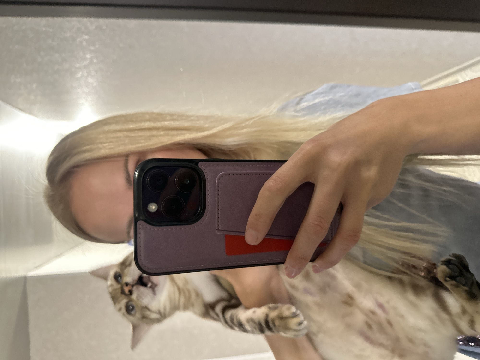 A woman with long blonde hair is taking a mirror selfie. She is holding a cat, which is looking up and to the left with its mouth open. The woman is holding a smartphone with a purple case and a red card visible in a slot on the case. The background appears to be an indoor space, possibly a bathroom or hallway, with light-colored walls and a reflective surface, suggesting a mirror. The location is not discernible from the image itself, and thus, cannot be stated to be in Energetik, Russia.