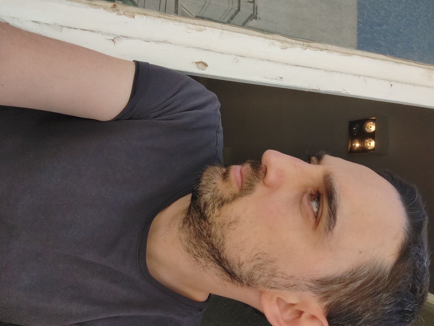 An eye-level profile shot captures a man's head and upper torso, facing towards the right side of the frame. He has short, dark hair with streaks of grey, a neatly trimmed beard and mustache, and light-colored eyes. He is wearing a plain, dark grey crew-neck t-shirt. His expression is neutral and contemplative, with his gaze directed forward.
In the background, positioned above the man's shoulder, is a section of a window frame with extensively peeling and chipped white paint, suggesting an older or distressed architectural element. Below this frame, the background recedes into a darker interior space. On a dark wall to the right of the man's head, there is a square wall-mounted light fixture. It features four circular lightbulbs, which are illuminated and casting a warm, yellowish glow. The overall lighting suggests an indoor setting, possibly during evening hours or in a dimly lit room. No specific activity is depicted beyond the man's steady gaze. There is no visible text in the image. While the location context is Tel-Aviv, Israel, no specific visual cues in the image confirm this.