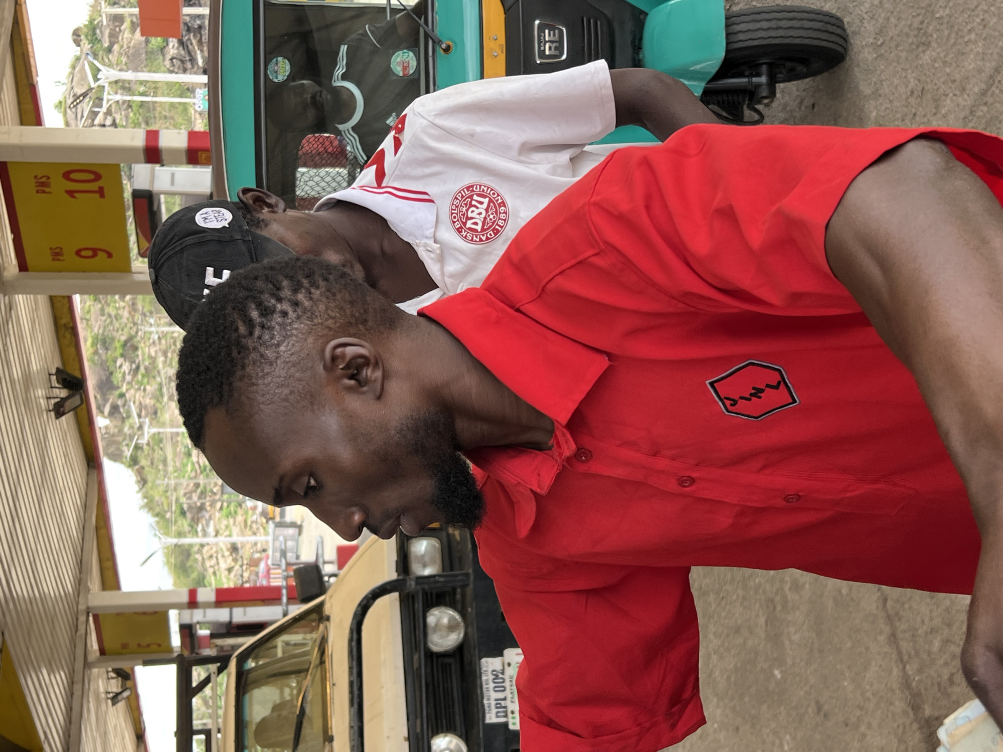 The image depicts two men near a gas station in Gura Top, Nigeria. One man is wearing a red shirt with a black logo on the pocket. He has a beard and short, cropped hair. He is looking down. Another man, wearing a white shirt with a Danish soccer logo and a black baseball cap, stands behind him. The men are next to a vehicle, possibly a tricycle, painted in green and black. In the background, there's a fuel price sign showing "PMS 9" and "PMS 10." Another vehicle with license plate "DPL 002" is visible. The environment appears to be a commercial area, given the gas station and vehicles.