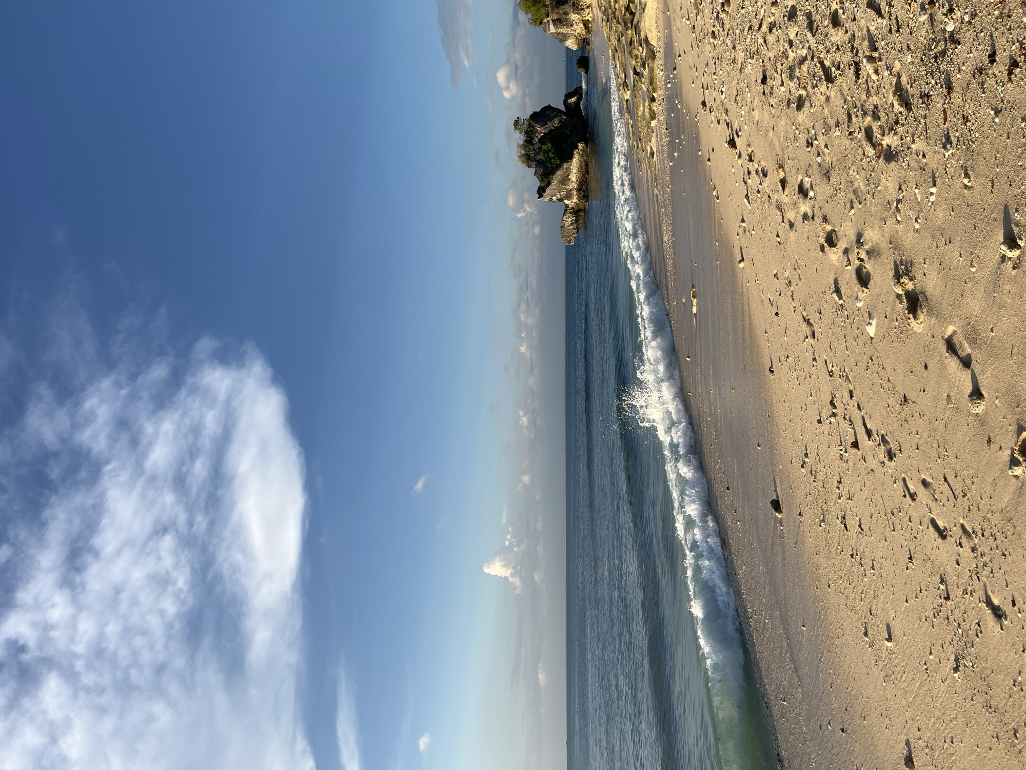 The image depicts a beach in Kuta Selatan, Indonesia. The sky is blue with scattered white clouds. The ocean is visible, with waves breaking on the sandy shore. The beach is light brown and has scattered stones and footprints. A rocky outcrop with vegetation is seen in the distance along the coastline.