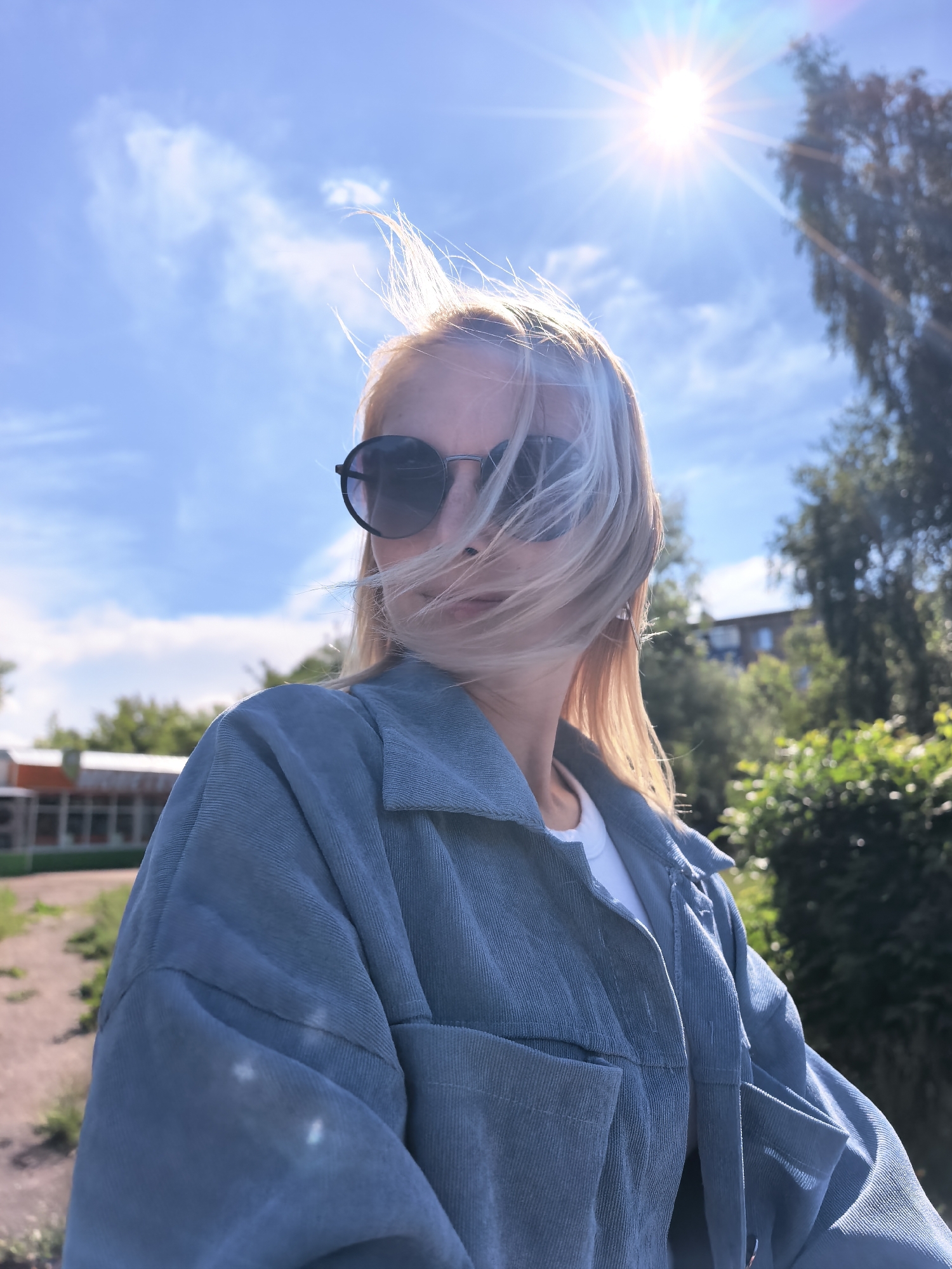 The image displays an adult female outdoors on a bright, sunny day. She has blonde hair, which is windswept and partially obscures her face and dark, round sunglasses. She is wearing a light blue or teal corduroy-textured jacket over a white t-shirt. The sun is prominently featured in the upper right quadrant of the image, creating significant lens flares and backlighting her hair, giving it a bright, luminous edge. The sky is clear blue with sparse white clouds. In the mid-ground to the left, a building with a reddish-orange sloped roof and multiple windows is visible, partially behind a fence or railing. To the right, green trees and bushes partially obscure another multi-story building. A dirt or unpaved area is visible in the lower-left foreground. The specific location as Sterlitamak, Russia, is not identifiable from visual elements within the image.