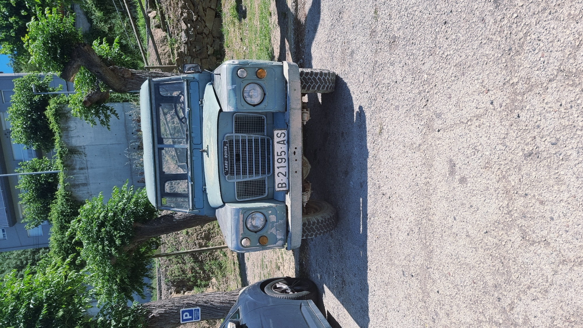 A light blue Land Rover is parked on a gravel surface in Montagut i Oix, Spain. The vehicle is facing forward and is positioned centrally in the frame. The Land Rover has a classic design with a prominent grille and round headlights. A license plate with the characters "B-2195-AS" is attached to the front bumper. To the left of the Land Rover, a dark-colored car is partially visible. In the background, there are trees with lush green foliage and a concrete structure, possibly a building, with some stone walls and fencing. The scene appears to be outdoors on a sunny day, with shadows cast on the ground.