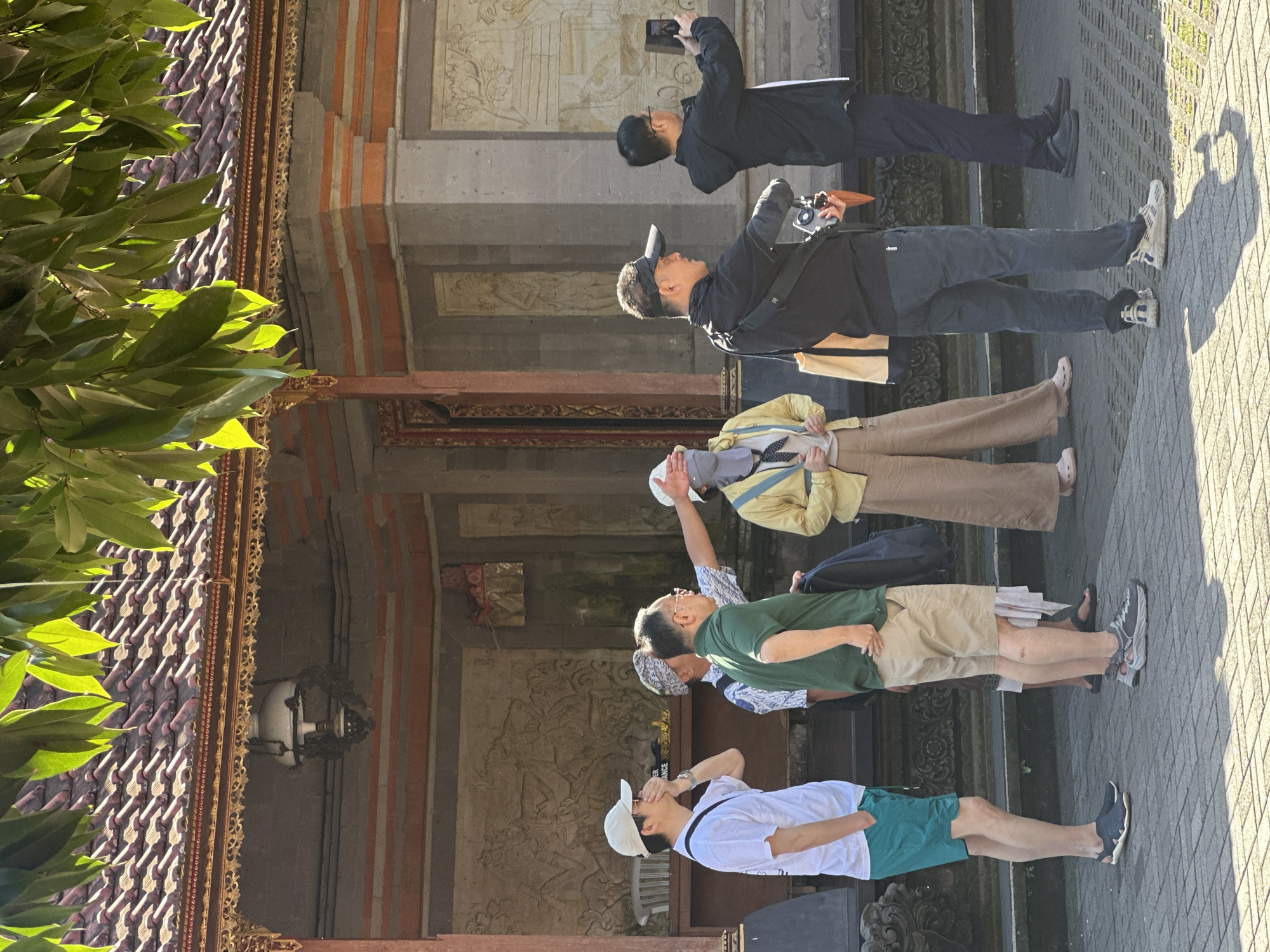 The image shows a group of tourists standing in front of a Balinese temple building, likely in the city of Ubud District, Indonesia, based on the architectural style and tropical foliage. The building features a traditional tiled roof, ornate eaves, and stone carvings on its walls.
In the foreground, several individuals are visible from the waist down, standing on a paved walkway. From left to right, the first person wears teal shorts and a white t-shirt. Next to them, a person in khaki shorts and a green t-shirt has one hand behind their back and the other pointing towards the temple. Behind them, a person in a patterned shirt is also visible. Further right, a person in tan pants and a yellow jacket appears to be looking at their phone, while another person in dark pants and a black jacket holds a camera. Another individual in dark pants and a black jacket is on the far right, taking a photo with a mobile phone.
The environment is brightly lit by sunlight, casting shadows on the pavement. Lush green leaves from an overhanging tree are in the upper portion of the frame, partially obscuring the roof of the temple. The overall scene suggests a cultural or historical site visit.