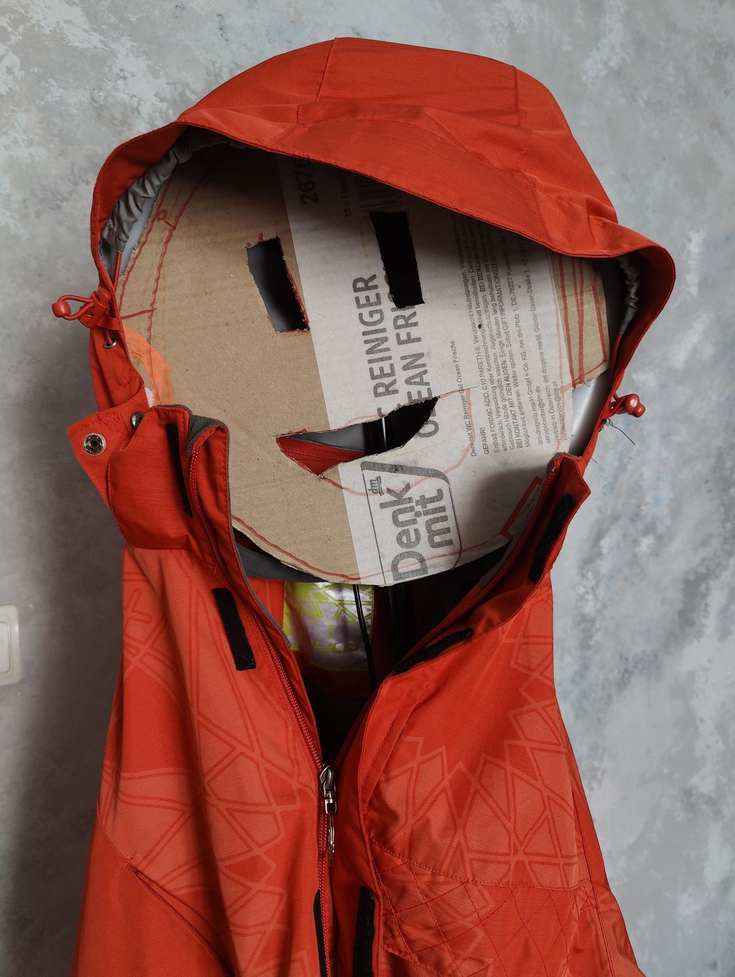 A brightly colored orange hooded jacket is hung on a wall, obscuring a cardboard cutout. The cardboard has been fashioned into a mask-like shape with two eyeholes and a mouth opening. Text is visible on the cardboard, including "REINIGER" and "Ocean Frische," suggesting it may be from a cleaning product. The jacket has a zipper that is partially open, revealing a glimpse of what appears to be yellow packaging inside. The wall behind the jacket has a textured, gray appearance, resembling concrete or stucco. The scene is likely indoors, given the wall texture and the way the jacket is displayed. There is no discernible information in the image that specifically places the scene in Yarmolyntsi, Ukraine.