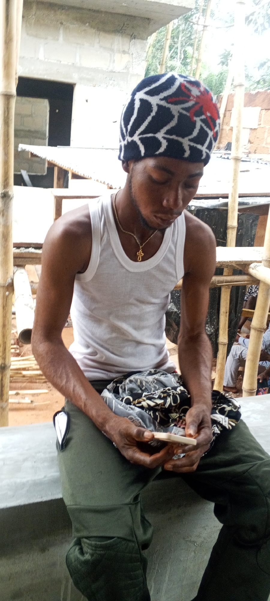 A man is sitting outside and looking at a rectangular object he is holding. The man is wearing a white tank top, a gold cross necklace, dark green pants, and a navy blue and white knit cap with a red design on top. The man's arms and upper chest are visible, and his skin is brown. On his lap, there is a pile of black and white patterned cloth. The man is sitting on a gray surface.
In the background are bamboo poles and a partly constructed concrete structure with a square opening. Other people are partially visible behind the bamboo poles. The location is likely in the city of Unknown, Unknown.