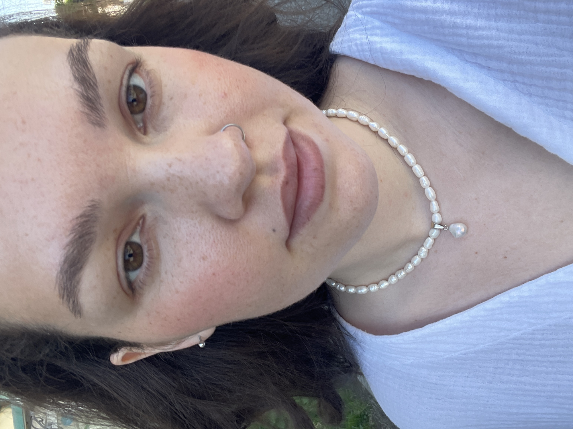 A close-up, tilted selfie shows a woman with brown hair, brown eyes, and a nose ring. She wears a white, textured shirt and a pearl necklace with a pearl pendant. Her skin tone is fair with freckles. The background is blurred and indistinct, showing glimpses of greenery. It is impossible to determine the location as Izhevsk, Russia based on the provided image.