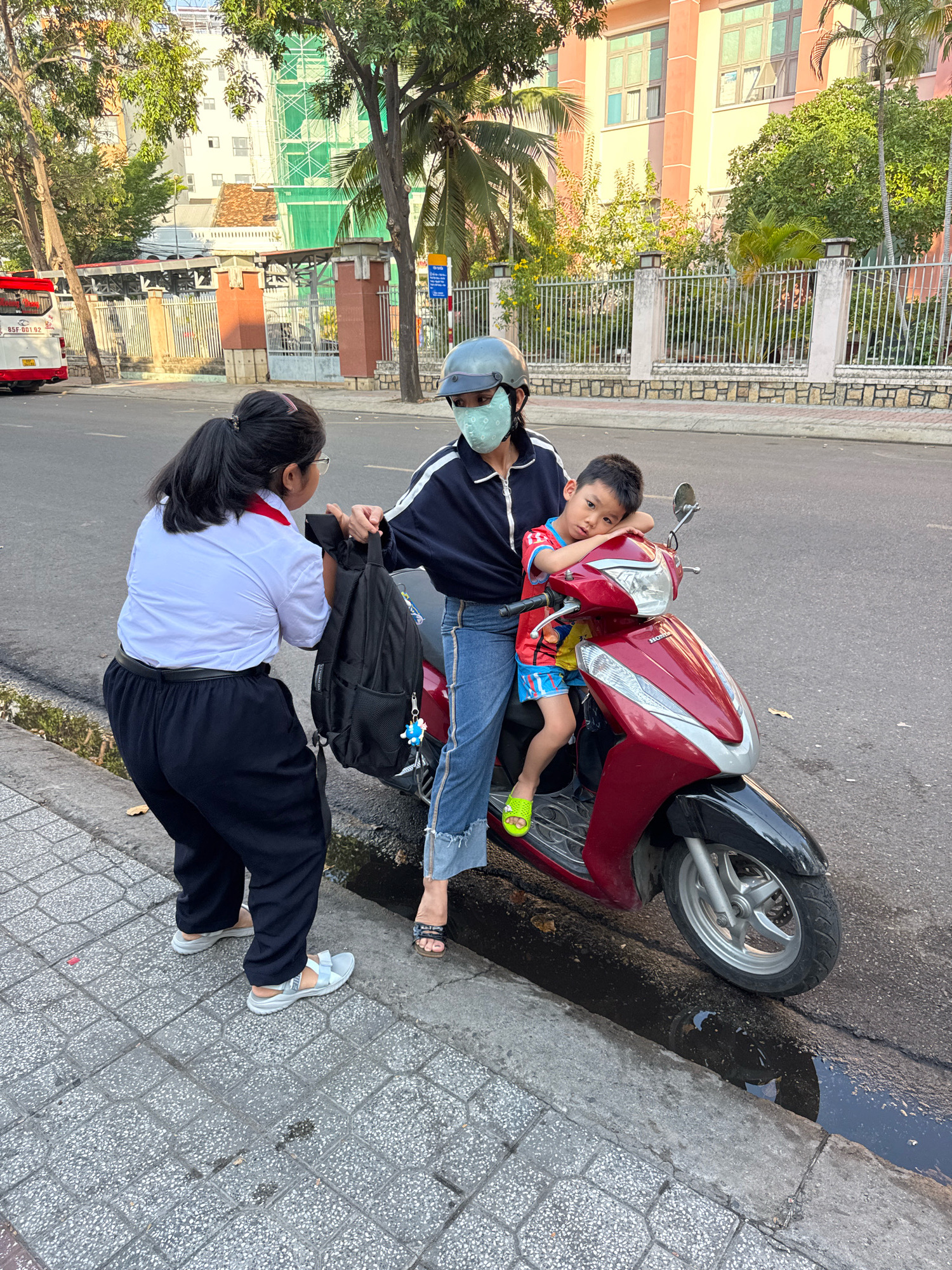 In Nha Trang, Vietnam, on a sunny day, a young girl in a white shirt, dark pants, and a red collar stands on a sidewalk next to a street, handing a black backpack to a woman wearing a helmet, a face mask, a dark jacket, and blue jeans, who is sitting on a red motorcycle. A young boy, wearing a colorful shirt and shorts, sits in front of the woman on the motorcycle, with his head resting on her arm. The motorcycle is parked on the side of the road, which has a drainage ditch filled with dark water. In the background, there are palm trees, buildings under construction with green netting, a fence, and a bus. The scene appears to be in front of a school or residential area.