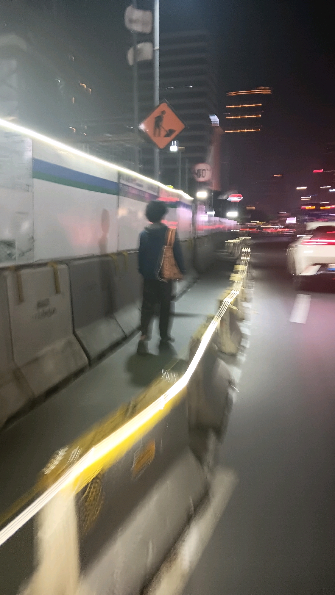 The image depicts a person walking along a sidewalk at night in Central Jakarta, Indonesia. The scene is characterized by motion blur, suggesting the photograph was taken from a moving vehicle or with a slow shutter speed. The person, seen from behind, is wearing a dark jacket and pants and carrying a woven bag over their shoulder. They are walking on a raised pedestrian walkway bordered by concrete barriers. To the right of the walkway, a street is visible with moving vehicle lights, including a white car. Overhead, construction signage, including a warning sign with a silhouette of a worker, is present, indicating ongoing work in the area. The background shows blurred city lights and buildings. The overall atmosphere is nocturnal and urban, with the motion blur contributing to a dynamic and somewhat abstract representation of the environment.