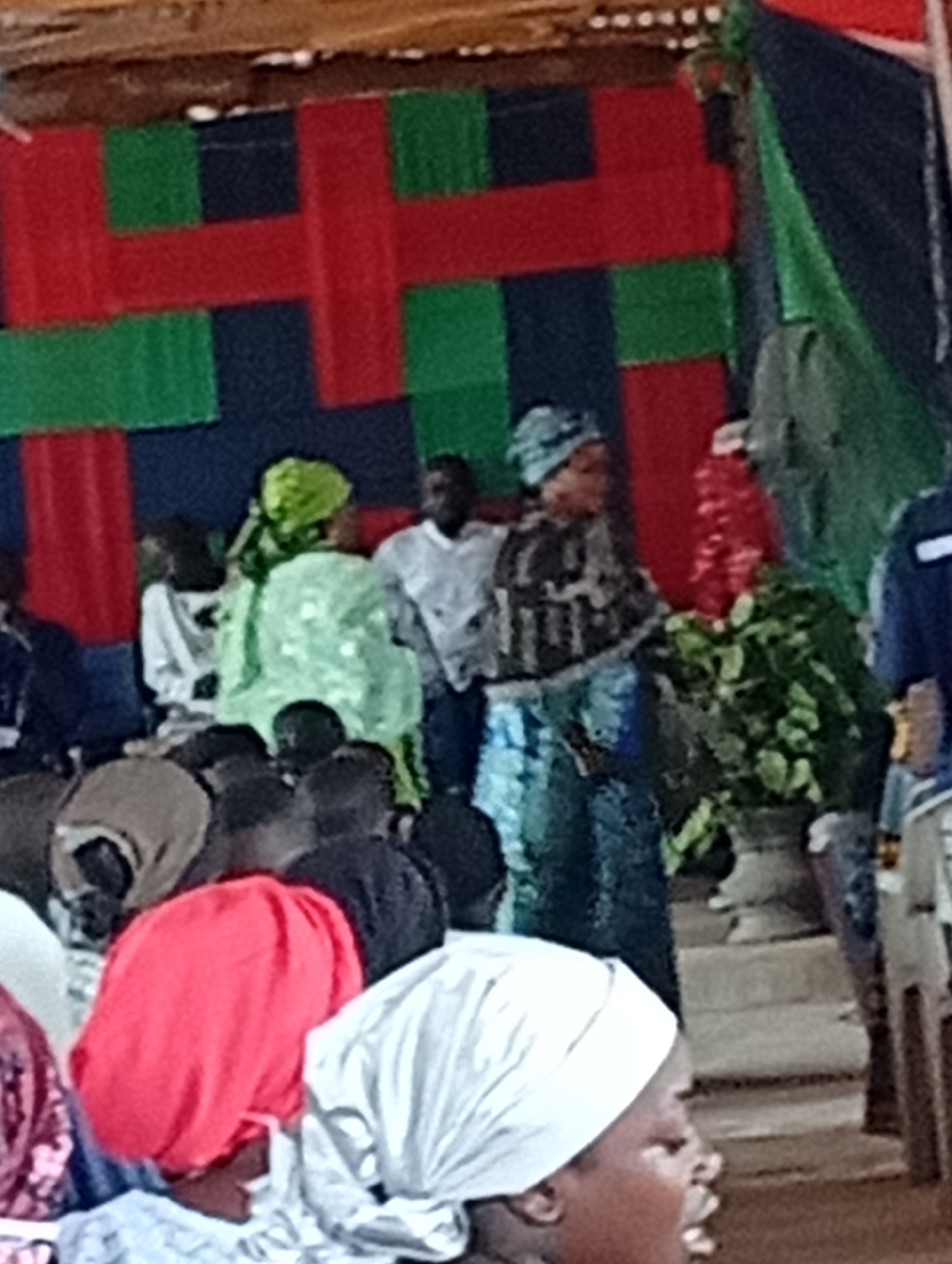 The image depicts a gathering of people, with some seated and others standing. The background features a decorative arrangement of red and green fabric panels, creating a visually striking backdrop. Several individuals are wearing head coverings in vibrant colors like red, green, and white. One woman in the foreground is wearing a blue and black patterned garment and a blue head covering. Another woman, positioned slightly behind her, is dressed in a light green outfit and headscarf. In the middle ground, a man in a white shirt is visible. A potted plant with lush green leaves stands to the right of the group. The overall scene suggests an event or a public gathering, possibly a cultural or religious ceremony.