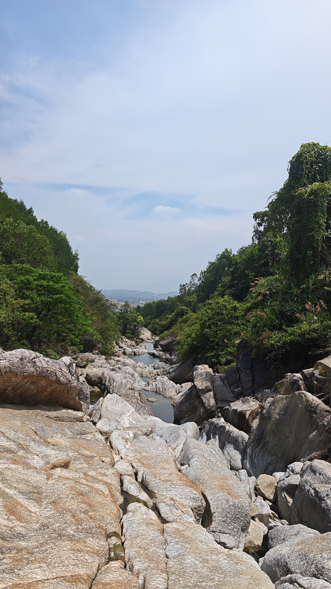 The image captures a rocky, mountainous river scene in Đà Nẵng, Vietnam. In the foreground, there are large, weathered rocks, primarily of light brown and grey color. Water flows through channels between the rocks. Lush green vegetation covers the hillsides on either side of the water. A faint view of the city can be seen in the distance beneath a blue sky with some scattered clouds. The environment appears natural, untouched, and dominated by rocky terrain and dense vegetation.