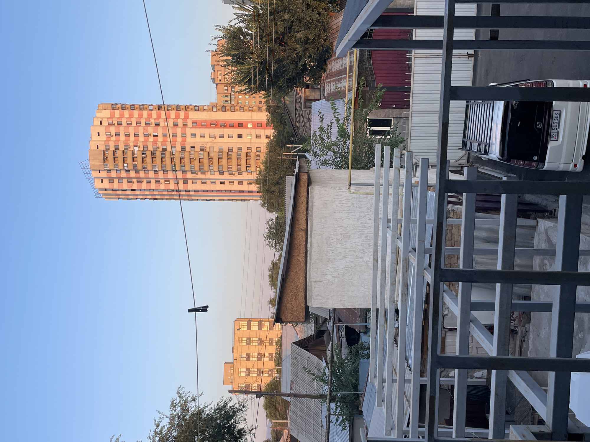 This image captures a view of a residential area in Almaty, Kazakhstan, under a clear, pale blue sky. In the background, a tall, light-colored apartment building with a pattern of pink and beige rectangles dominates the skyline. To its left, a shorter, beige building with evenly spaced windows is visible. The foreground is filled with a cluster of smaller, older-looking buildings with corrugated metal roofs, some appearing weathered and dilapidated. A white minivan with a roof rack is parked in a driveway in the lower right portion of the frame. A clothes peg hangs from a wire stretched across the middle ground. The scene appears to be a typical urban or semi-urban environment with a mix of modern and older structures.