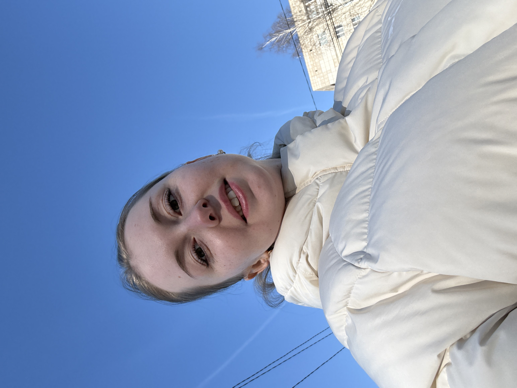 A woman is looking down at the camera. The background is a clear blue sky, with a building visible in the upper right. She is wearing a white puffer jacket. The photo appears to be taken from a low angle, tilted. Electric wires are also visible against the sky.