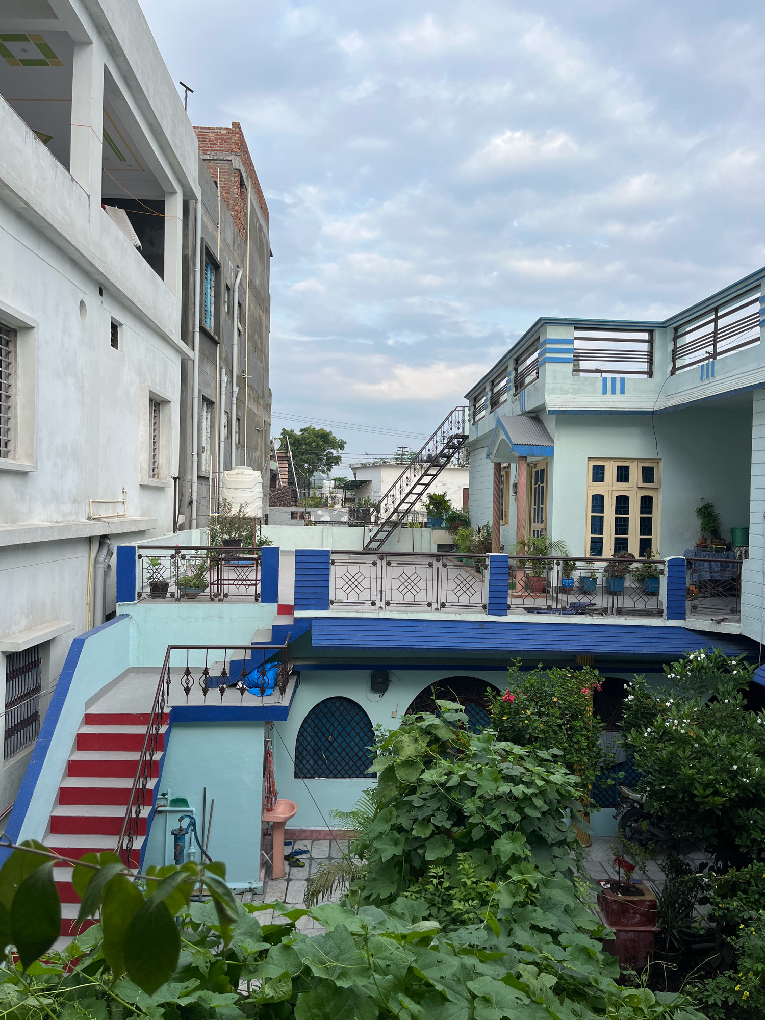 The image depicts a residential area in Rishikesh, India, under a cloudy sky. The scene is dominated by multi-story buildings with varying architectural styles and colors.
On the left, a white building with grey trim and multiple windows is partially visible. Adjacent to it is a building under construction, with exposed brickwork at the top and grey plaster on the lower levels. Plumbing pipes are visible on the exterior of this building.
The central and right portions of the image showcase a light blue and white two-story house. This house features balconies with decorative railings, an external metal staircase leading to the upper level, and multiple windows with wooden frames. Potted plants are arranged on the balconies and terraces. A prominent water tank is situated on the roof of the adjacent building.
In the foreground, lush green foliage and plants are visible, some with red and white flowers, suggesting a garden or overgrown vegetation. The ground level appears to be paved with stone tiles. A pink basin and a blue plastic mat are visible near a blue-colored wall. A bicycle or motorcycle is parked in the background. The overall impression is of a densely populated urban residential environment.