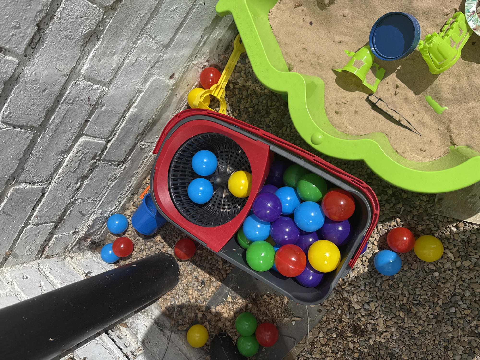 The image depicts a collection of colorful plastic balls, primarily red, blue, yellow, and green, scattered across gravel and sand. The balls are partially contained within a red and grey plastic mop bucket, which appears to be used for storage. Adjacent to the bucket is a bright green plastic sandbox, also containing sand and a few green plastic toys, including a small shovel and what looks like a mold. A black downpipe is visible on the left side of the image, situated next to a white brick wall. The scene is outdoors and bathed in bright sunlight, casting distinct shadows. The location is stated to be Darlington, United Kingdom. There are no people or animals visible in the image.