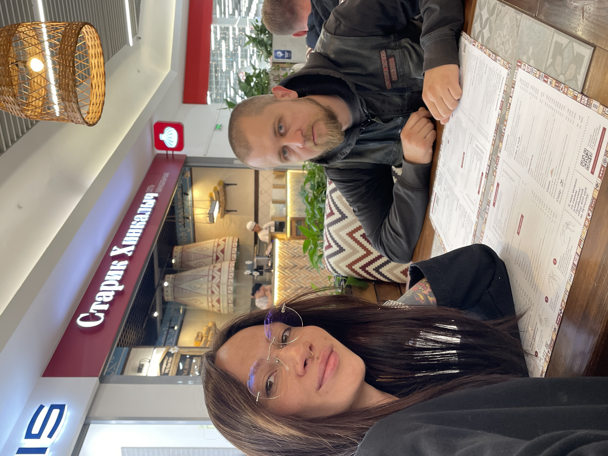 This image captures a selfie-style shot of two individuals seated at a wooden table in a "Старик Хинкалыч" (Old Man Khinkalych) restaurant, located within the Greenwich (ТРЦ Гринвич) shopping mall in Yekaterinburg, Russia.
The foreground is dominated by a woman on the left, who is taking the photograph. She has long, dark brown hair, parted on the side, and is wearing rimless circular eyeglasses with a slight blue glare on the lenses. She has a small stud piercing on her right nostril and a septum ring. Her lips are slightly parted in a subtle smile. She is dressed in a black long-sleeved top, and a colorful tattoo is partially visible on her right forearm.
Opposite her, on the right side of the frame, sits a man with a closely-shaved head, blue eyes, and a light brown goatee and mustache. He wears a dark grey hooded sweatshirt beneath a black sleeveless vest. The vest features a white patch on the left chest area, identifying "НОЧНЫЕ ВОЛКИ" (Night Wolves, a motorcycle club) and "ИЖЕВСК" (Izhevsk). His hands are resting on the table in front of him.
Two open menus, printed in Cyrillic (Russian), are visible on the wooden table between the two individuals. The menus list various items, including "Хинкали" (Khinkali), "Кофе и чай" (Coffee and Tea), and "Пиво" (Beer). The table is covered in sections with a patterned grey and white surface.
The background shows the interior of the "Старик Хинкалыч" restaurant. A prominent red sign with white Cyrillic lettering "Старик Хинкалыч" is visible, along with a smaller red square sign featuring a white khinkali dumpling icon. The restaurant's decor includes large, light-colored conical structures with geometric patterns. Potted green plants are placed around the seating area. The upholstered booth seating features a beige, brown, and white chevron pattern. In the upper right corner, a large wicker pendant lamp hangs from the white ceiling, which is fitted with recessed lighting. In the far background, behind the man, kitchen staff in white uniforms are partially visible. To the upper left, a blue-lit, stylized "IS" sign, part of the "GREENWICH" mall logo, is visible against a white background.