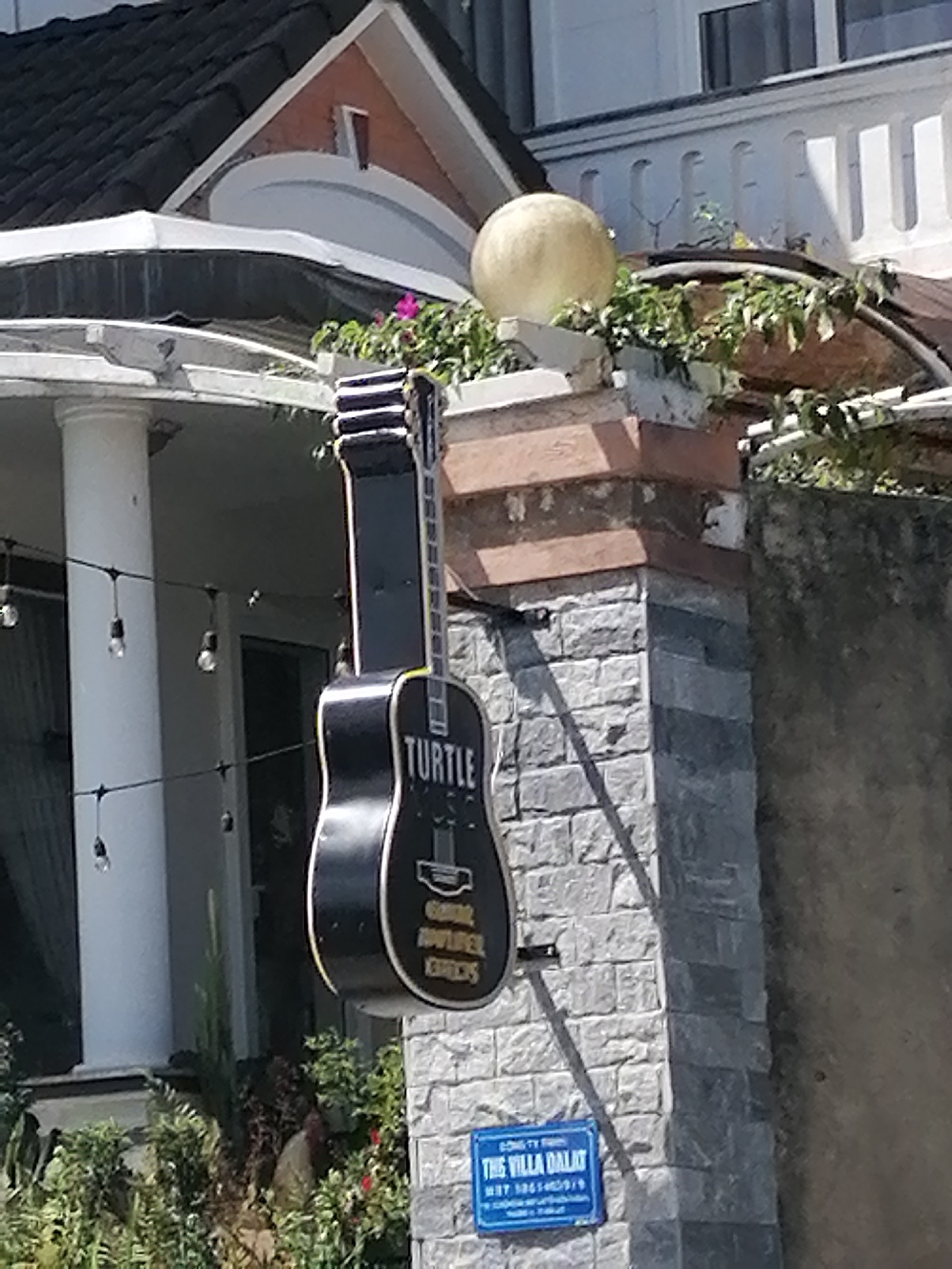 The image shows a black guitar-shaped sign hanging on a stone pillar in front of a building. The sign has "TURTLE" written on it in white letters, along with other text that is partially legible. Below the guitar sign, there is a blue rectangular sign with "THE VILLA DALAT" written in white. The building appears to be a hotel or guesthouse, possibly with a restaurant or bar, given the guitar motif. The environment is outdoors, with some greenery and string lights visible. The lighting suggests it is daytime. The architectural style of the building, with its tiled roof and white columns, is consistent with many establishments in Da Lat, Vietnam, a city known for its French colonial architecture and scenic beauty. The sign itself is a common form of advertisement for businesses in tourist areas.