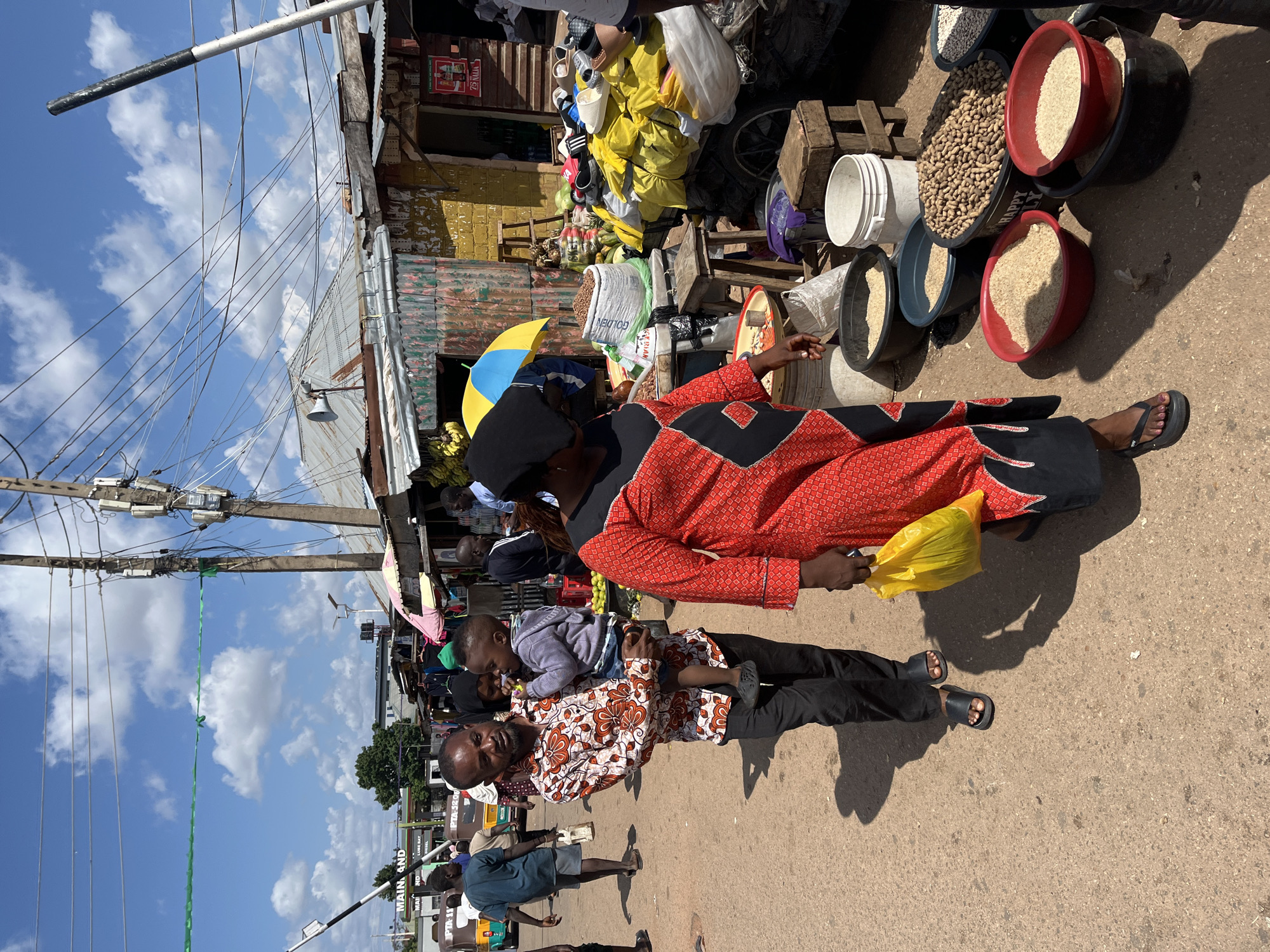In Gura Top, Nigeria, a man carries a toddler in his arms while walking down a dusty street lined with market stalls. The man wears a colorful patterned shirt and black pants, while the child is in a gray hoodie and blue jeans. Behind them, a woman in a red and black patterned dress walks with a yellow plastic bag. The market stalls display various goods, including bags of grains, nuts, and produce. Overhead, a tangle of electrical wires crisscrosses the sky, supported by utility poles. The scene is illuminated by bright sunlight, casting long shadows on the ground. Several other individuals are visible in the background, engaged in market activities.