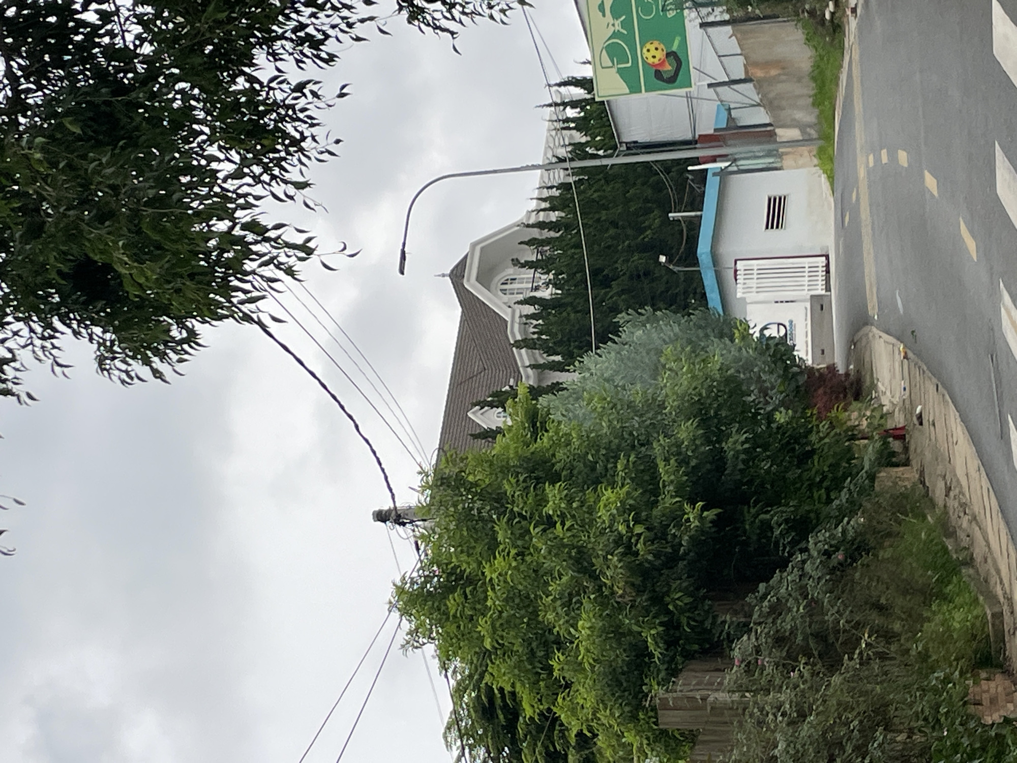 A cloudy day in Da Lat, Vietnam, is captured in this image. A grey, paved road curves upwards and to the right, marked with yellow dashed lines. On the left, a dense green tree and a wooden fence border the road. Further up the road, a multi-story white house with a dark roof and dormer windows is visible, surrounded by tall green cypress trees. A grey lamppost stands prominently in front of the house. To the right of the house, a white building with a light blue roof and a white gate is situated. A green sign with a yellow ball and some Vietnamese text is attached to a fence on the far right. The overall scene depicts a residential area on an overcast day.