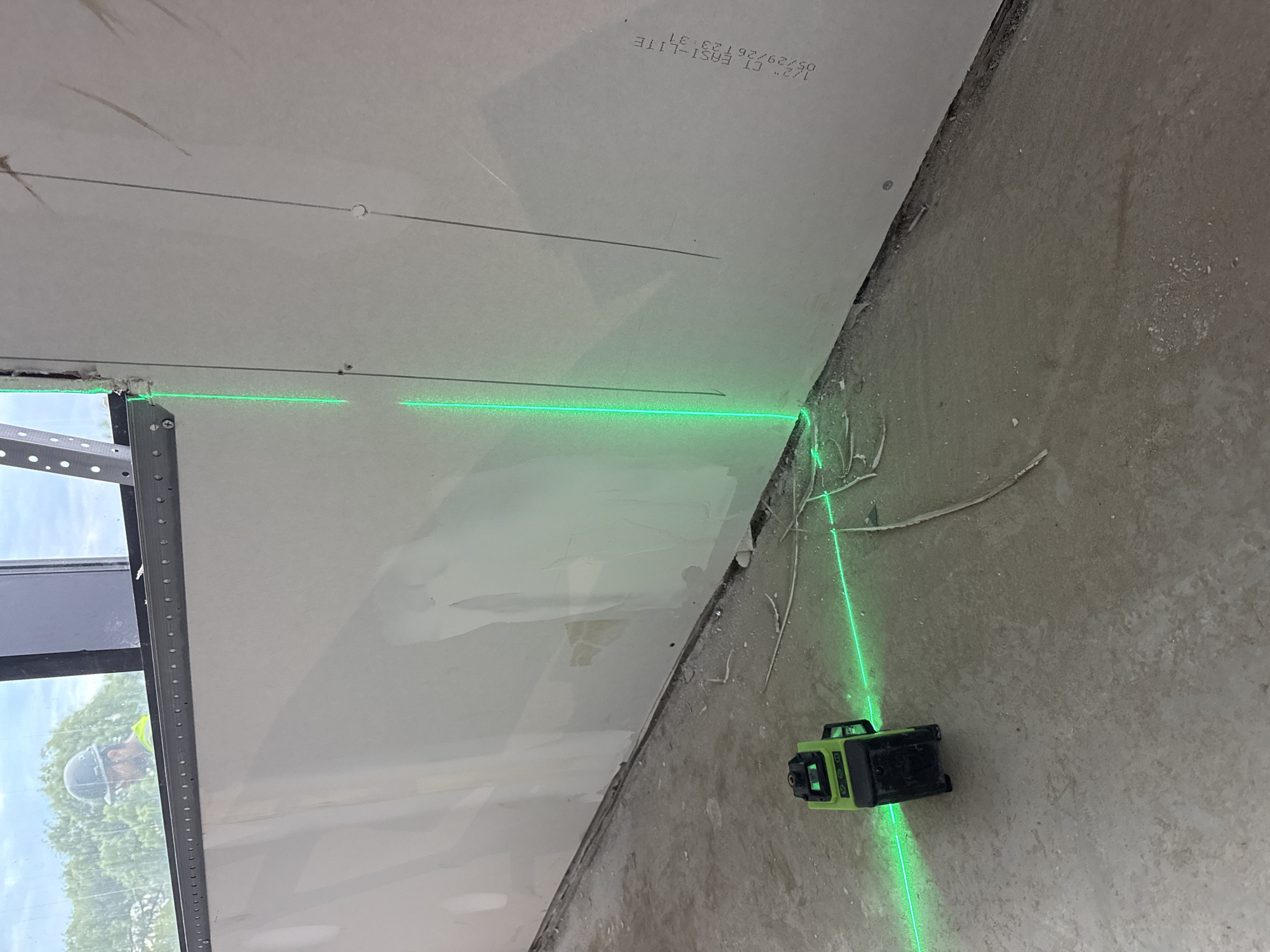 A green laser level is set up on a concrete floor, projecting a laser line. The laser is positioned in front of a wall constructed of drywall, which appears to be under construction or renovation. The wall has metal framing visible at the top and is marked with pencil lines. A worker wearing a hard hat and a high-visibility vest is visible through a window in the background, suggesting an outdoor environment beyond the construction area. The scene is indoors, likely a commercial or industrial space in Brampton, Canada, based on the provided location context.