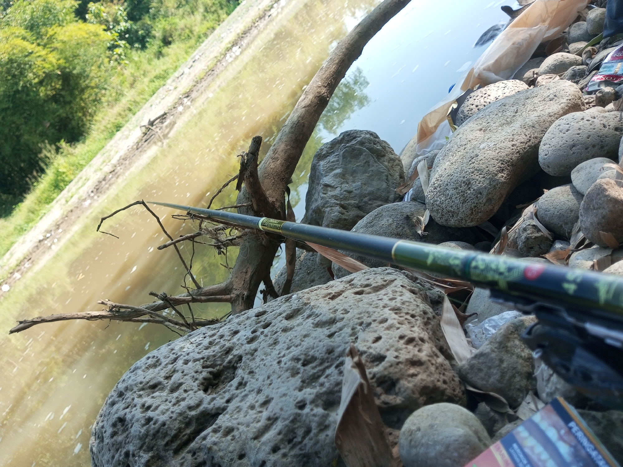 A close-up, angled shot shows a fishing rod resting on a rocky shore beside a body of water. The water, reflecting the green of the surrounding trees, appears murky and somewhat choppy with visible ripples. The shore is densely packed with rounded stones of varying sizes, many of which are porous and rough in texture. A weathered tree branch extends from the bank towards the water, its bare twigs reaching out. In the background, lush green foliage is visible, suggesting a natural, possibly rural environment. On the right side of the frame, partially obscured by rocks, are discarded items including a plastic bag and what appears to be packaging for cigarettes. The overall scene suggests a moment of calm by the water, with the fishing rod indicating the activity of angling. The location is identified as Wonosegoro, Indonesia, which is consistent with the tropical vegetation and type of river or lake depicted. There are no individuals present in the image, and no discernible actions other than the resting fishing rod.
