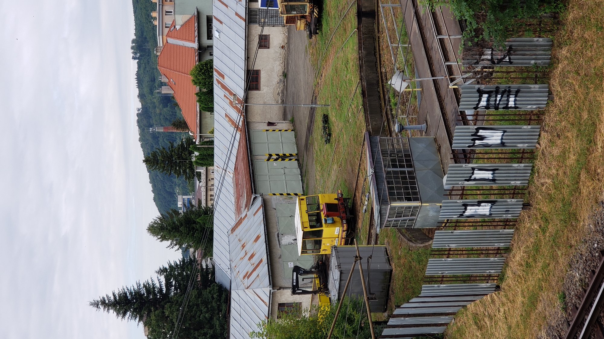 The image depicts an outdoor industrial area, possibly a tram depot or maintenance yard, located in Prešov, Slovakia. In the foreground, a corrugated metal fence with graffiti marks runs diagonally across the lower portion of the frame. Beyond the fence, a section of railway track is visible.
The central focus of the image is a yellow tram or rail vehicle parked on a track. This vehicle has large windows and appears to be a smaller, possibly specialized, unit. To the left of the tram, a piece of heavy machinery with a crane arm is parked. Several industrial buildings with corrugated metal roofs, some showing signs of rust, are visible in the background. These buildings appear to be garages or workshops, with large bay doors. The environment is somewhat overcast, suggesting a cloudy day. In the far background, a hilly, tree-covered landscape with some buildings on the slopes and a prominent red-bricked chimney can be seen. The overall scene conveys a functional, working industrial environment.