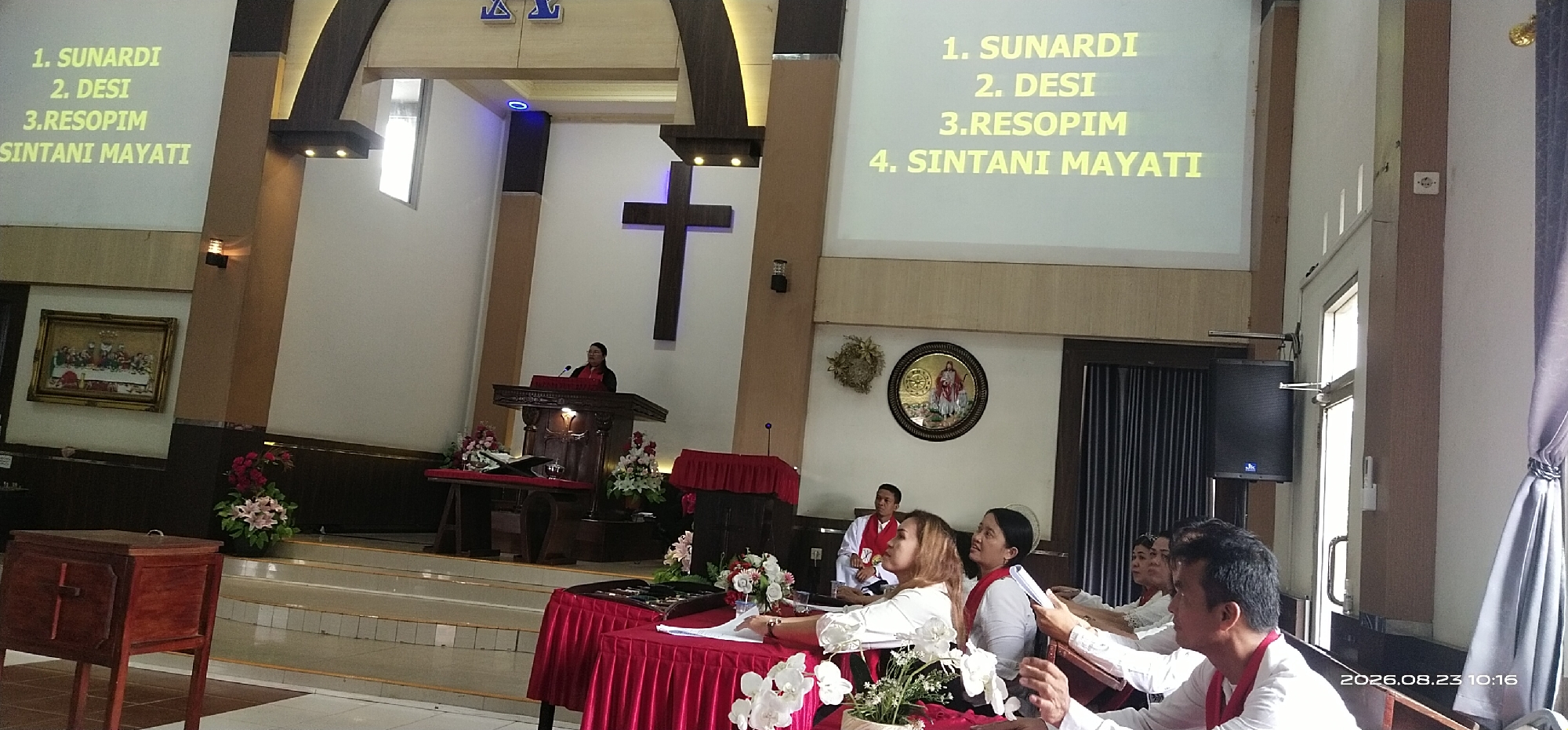 The image shows an interior view of a church, possibly in Jaar, Indonesia, as indicated by the timestamp which includes "2026.08.23 10:16". In the foreground, there are several individuals seated at tables covered with red cloths, some appearing to be engaged in a service or meeting. In the background, a person stands at a pulpit, addressing the congregation. A large wooden cross is prominently displayed on the wall behind the pulpit. A projector screen displays text, likely part of a presentation or worship service, with names and numbers. The church architecture features wooden beams and light-colored walls. The overall scene suggests a religious gathering.