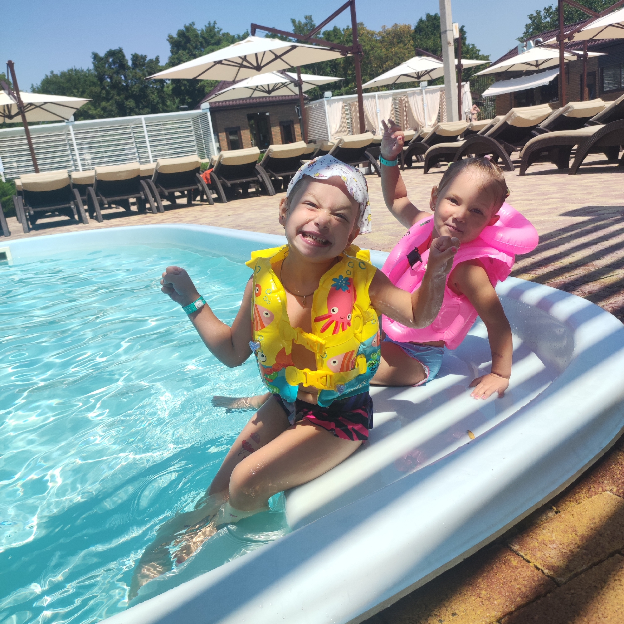 Two young girls are playing in a swimming pool. The girl in the foreground is wearing a yellow life jacket with cartoon octopus and fish designs and a headscarf. She is smiling broadly at the camera. The second girl, behind and to the right, is wearing a pink inflatable collar around her neck and a blue swimsuit. Both girls are in the water, which is clear and blue. The pool is bordered by a white rim. In the background, there are rows of empty lounge chairs under white umbrellas, suggesting a resort or hotel setting. The ground around the pool appears to be paved with brick-like tiles. The sky is clear and blue, indicating a sunny day. The image was taken outdoors.