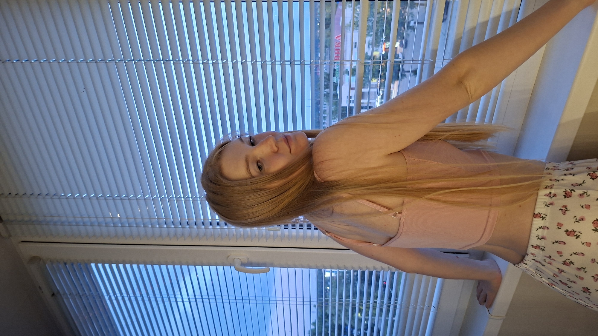 A young woman with long blonde hair is leaning on a windowsill, looking over her shoulder at the camera. She is wearing a pink tank top and white shorts with a floral pattern. She is standing in front of a window with blinds, which are partially closed, revealing a view of a city in the background. The lighting suggests it is either dusk or dawn. The overall scene is casual and domestic. The city in the background has been identified as Chaikovsky, Russia.