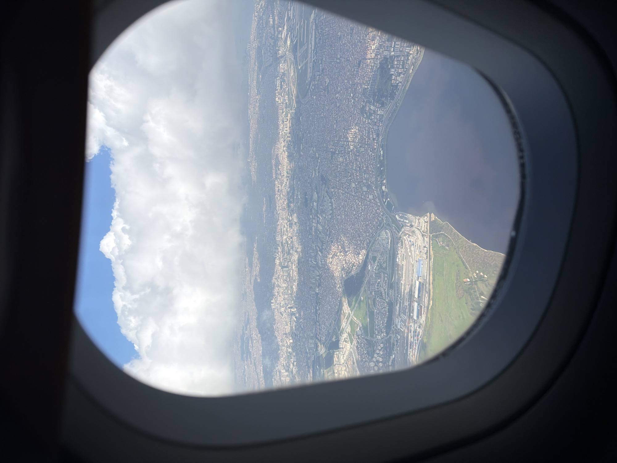 An aerial photograph, taken from the window of an airplane, captures a vast, densely populated coastal city under partial cloud cover during the daytime. The dark, rounded frame of the airplane window borders the image, framing the view.
The scene showcases an extensive urban landscape characterized by a multitude of buildings stretching along a prominent coastline. To the right of the image, a large body of dark blue water, likely a sea or ocean, extends to the horizon. To the left, a significant portion of the sky is visible, adorned with bright white cumulus clouds set against patches of clear blue sky.
Within the urban sprawl, a complex network of roads and infrastructure is discernible. A notable feature is a large industrial or port area, or potentially an airport, situated near the coast. This area is distinguished by expansive paved surfaces, numerous large structures, and what appear to be runways or cargo terminals. Interspersed throughout the city and near the coastal industrial zone are several green patches of land, possibly parks or undeveloped areas.
The photograph depicts a bright, clear day with visible sunlight, though large cloud formations are present. There are no people or visible text in the image. The perspective suggests the aircraft is at a relatively low altitude, offering a detailed view of the coastal city and its infrastructure.