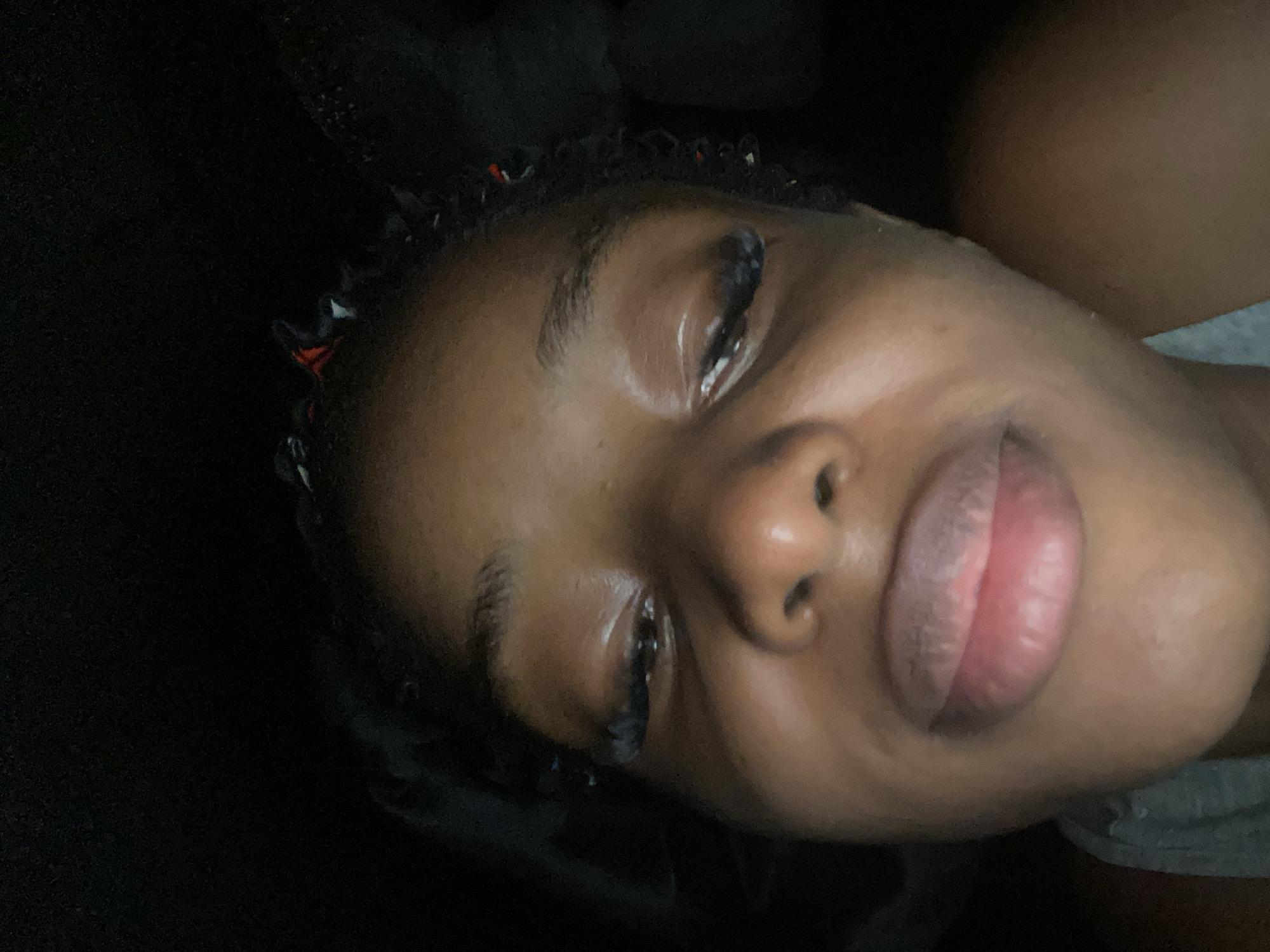 The image is a close-up selfie of a young Black woman with braided hair. She is looking downwards with her eyes partially closed and her mouth slightly open, revealing her lips. The lighting is dim, casting shadows on her face. She appears to be wearing a gray top. The background is dark and indistinct. The image does not provide any information about the location in Jos, Nigeria.