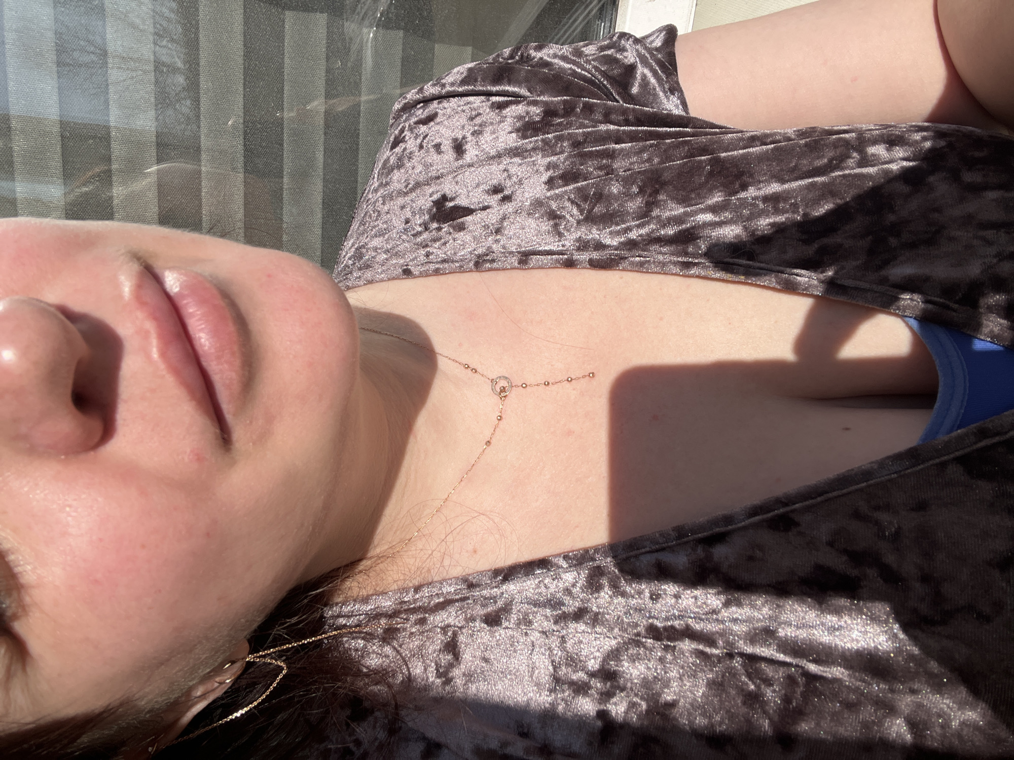A woman is seen from a low-angle perspective, lying down. Her face is partially visible, focusing on her lips and nose. She is wearing a brown, crushed velvet-like top. A delicate gold necklace adorns her chest. A glimpse of blue fabric is visible under the top, possibly underclothing. The background suggests a window or a partially translucent barrier with vertical lines, allowing sunlight to create stark shadows on her skin and clothing. There is no specific evidence in the image to suggest this photo was taken in the city of Noviy, Russia.