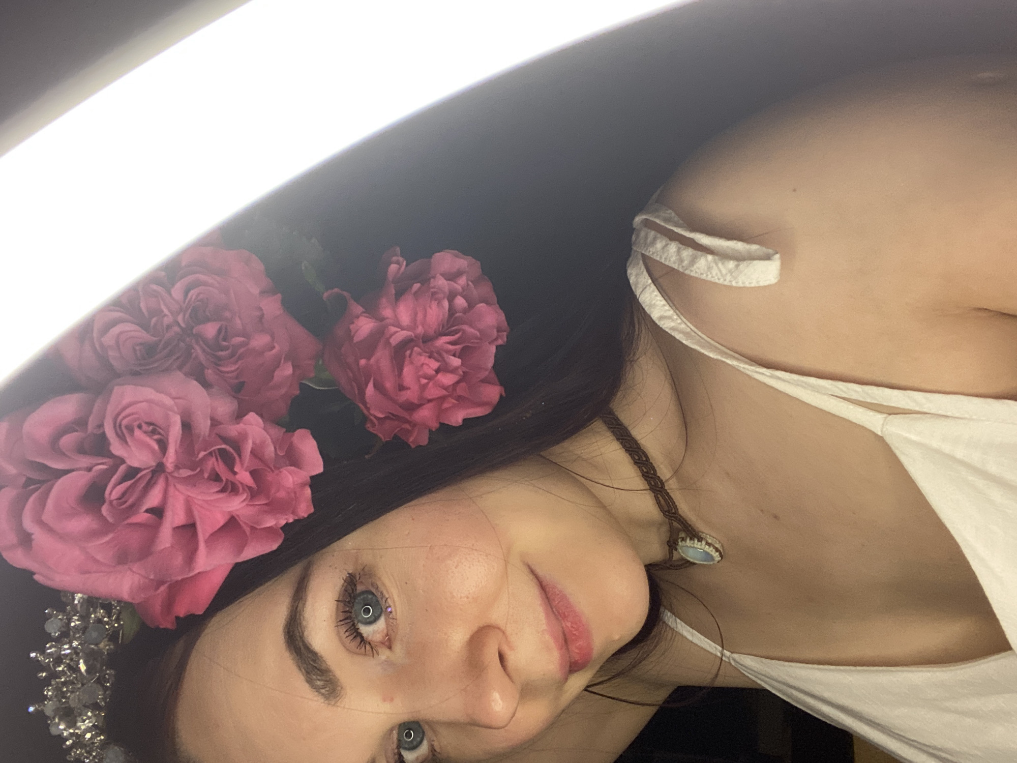 A close-up, tilted selfie features a woman with dark hair adorned with pink roses and a beaded headpiece. She wears a white tank top and a braided necklace with a pendant. The background is dark, with a bright light source to the left of the frame. There is no information indicating the location is Chaikovsky, Russia.