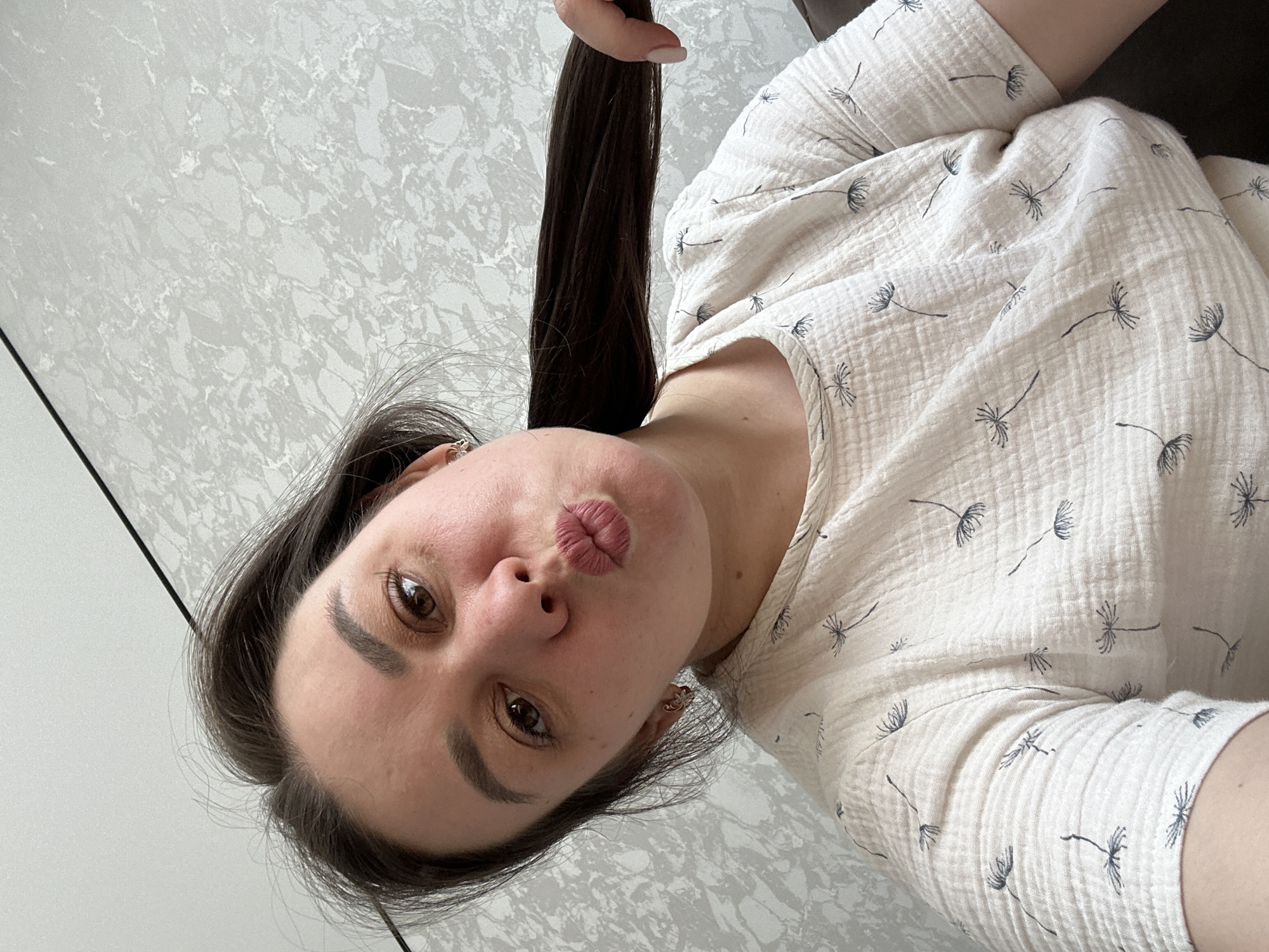 A woman is posing indoors, facing the camera at an angle. She is holding a section of her long, dark hair up with her hand. She has brown eyes, defined eyebrows, and is puckering her lips for the photo. She is wearing a light-colored top with a dandelion seed head pattern. The background is a light grey textured wall. There's no verifiable information about the location being Chaikovsky, Russia, in the image itself.